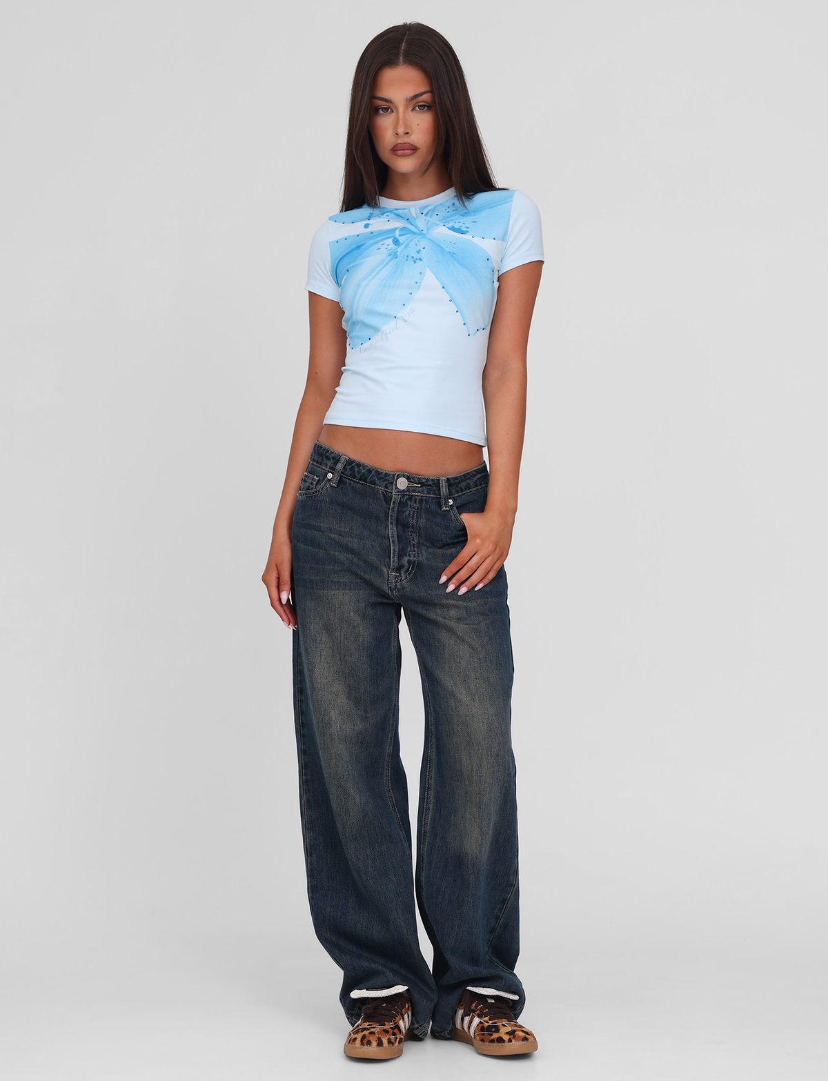 A woman with straight brown hair wears the TIGERMIST SAPHIRA TEE in blue, featuring a butterfly design and stretchy fabric, paired with baggy dark blue jeans and leopard print shoes, standing against a plain white background.
