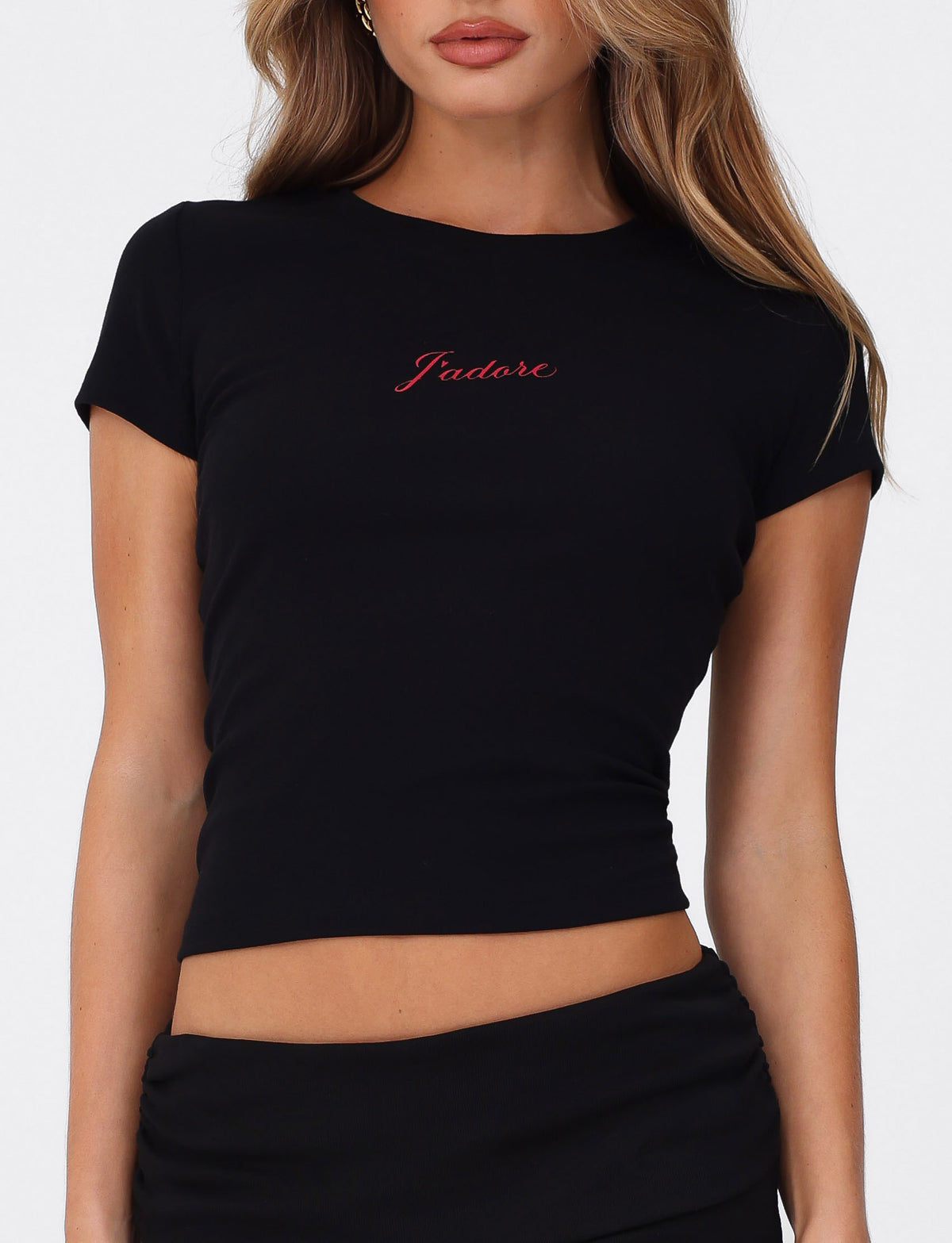 A woman wears the TIGERMIST SAPHIRA TEE in black, a fitted high-stretch top with a bold red J'Adore graphic on the chest, paired with a matching black skirt.