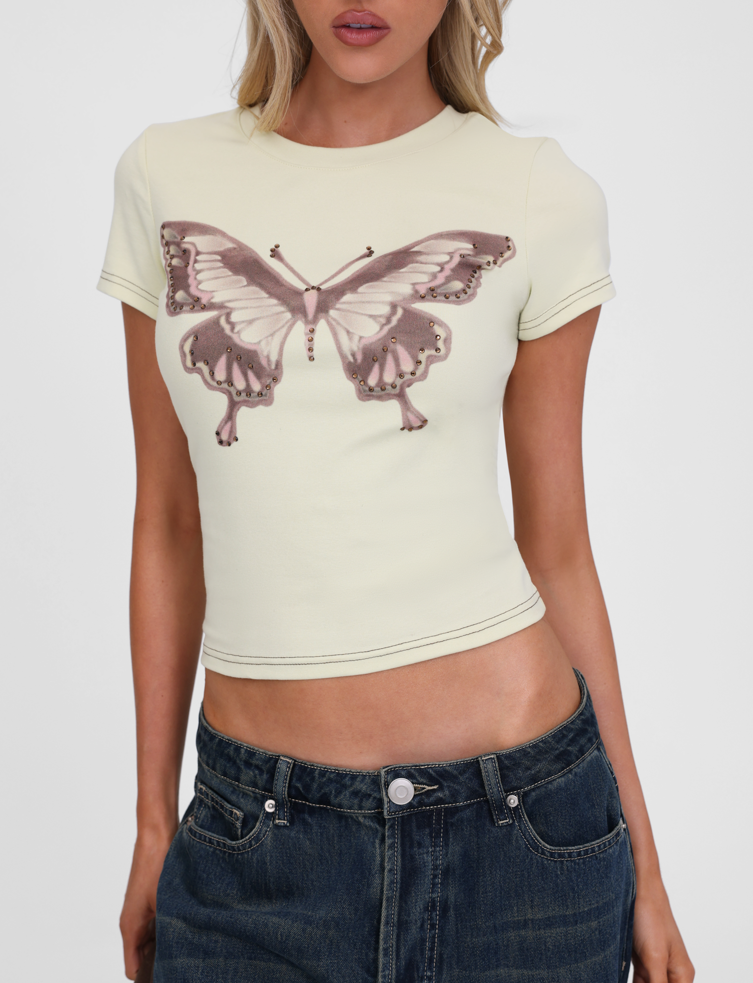 A woman wears the TIGERMIST SAPHIRA TEE in butterscotch—a fitted cotton top with cap sleeves and a large mauve butterfly graphic—paired with low-rise dark blue jeans. Her face is partially out of frame.