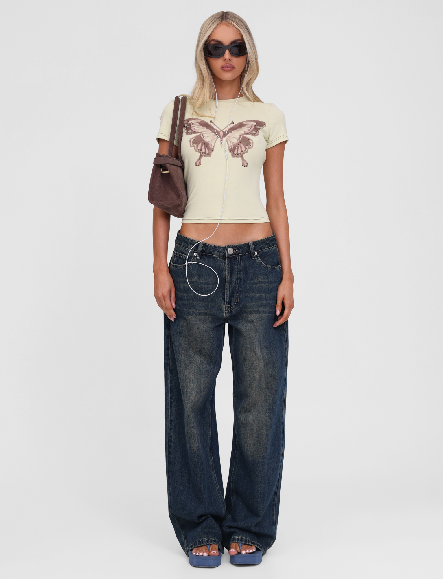 A woman wears TIGERMIST's SAPHIRA TEE in butterscotch with cap sleeves and a butterfly graphic, baggy blue jeans, blue sandals, sunglasses, a brown shoulder bag, and earphones hanging from her neck.