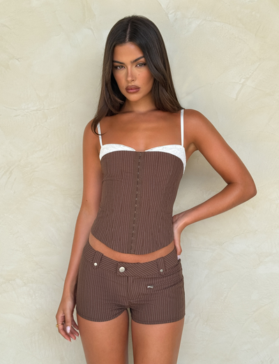 A woman with long brown hair wears the TIGERMIST ANTONELLA SHORT in chocolate pinstripe, paired with a matching corset top, standing against a beige textured wall and looking at the camera with a neutral expression.