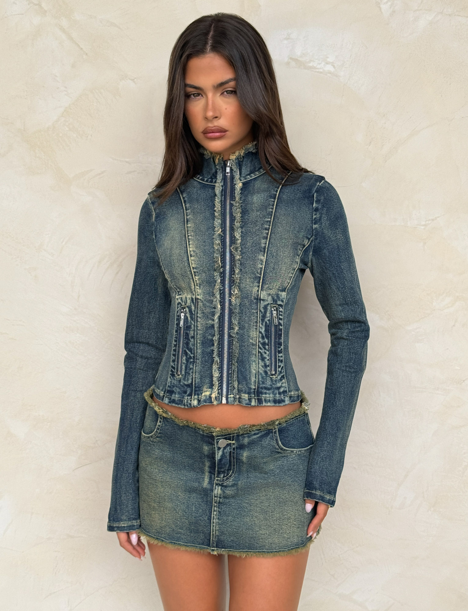 A woman with loose dark hair stands against a beige wall, wearing the TIGERMIST OPHELIE JACKET in denim blue with a high collar and matching mini skirt. Her expression is neutral.