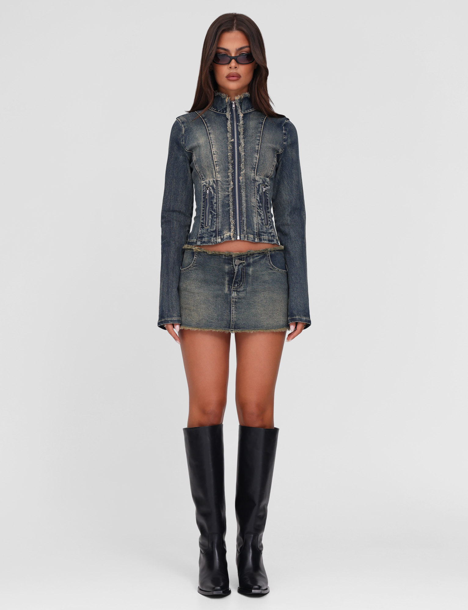 A woman stands facing forward against a white background, wearing the TIGERMIST OPHELIE JACKET in denim blue, paired with a matching mini skirt, black knee-high boots, and dark sunglasses.