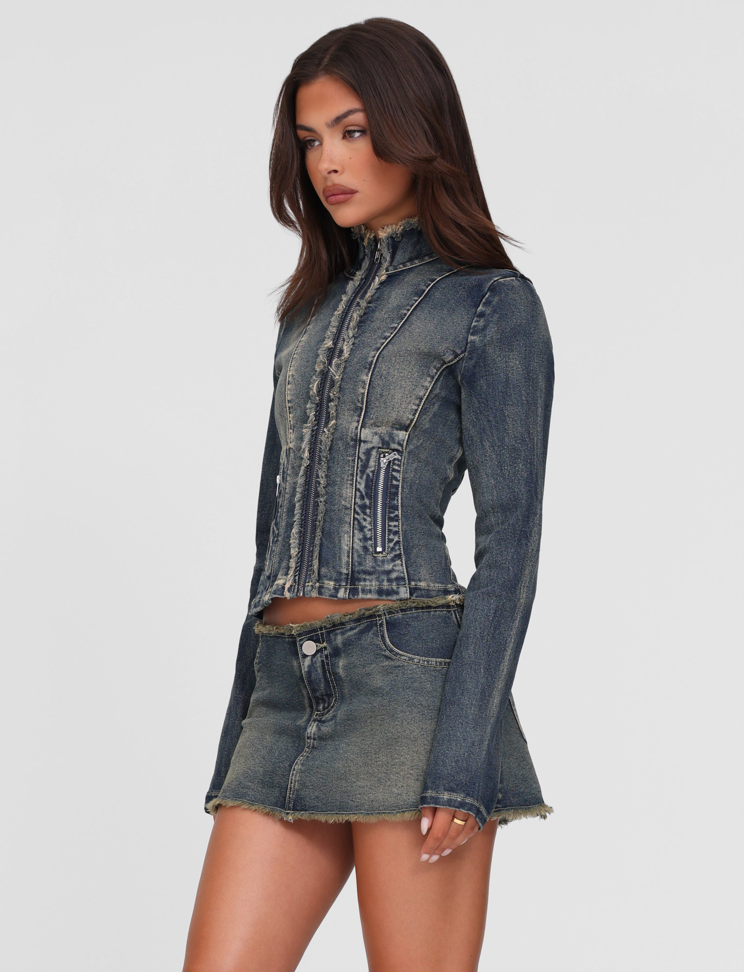 A woman models the TIGERMIST OPHELIE JACKET - BLUE, a fitted denim jacket in vintage blue with frayed edges, paired with a matching mini skirt, standing against a plain white background.