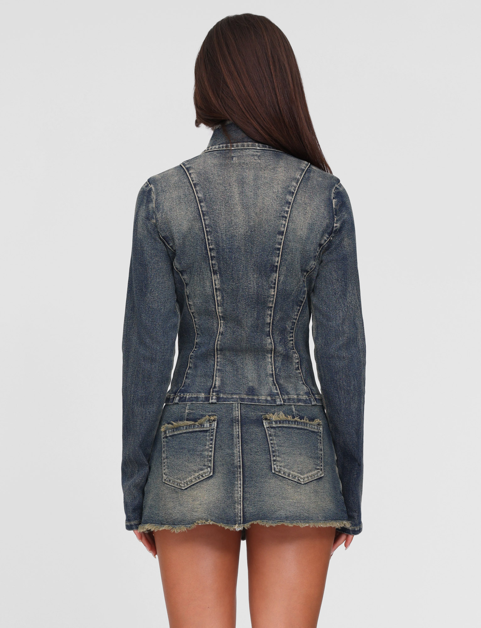 A woman with long brown hair, seen from the back against a plain light background, wears the TIGERMIST OPHELIE JACKET and matching short skirt in vintage wash blue denim with frayed edges and back pockets.