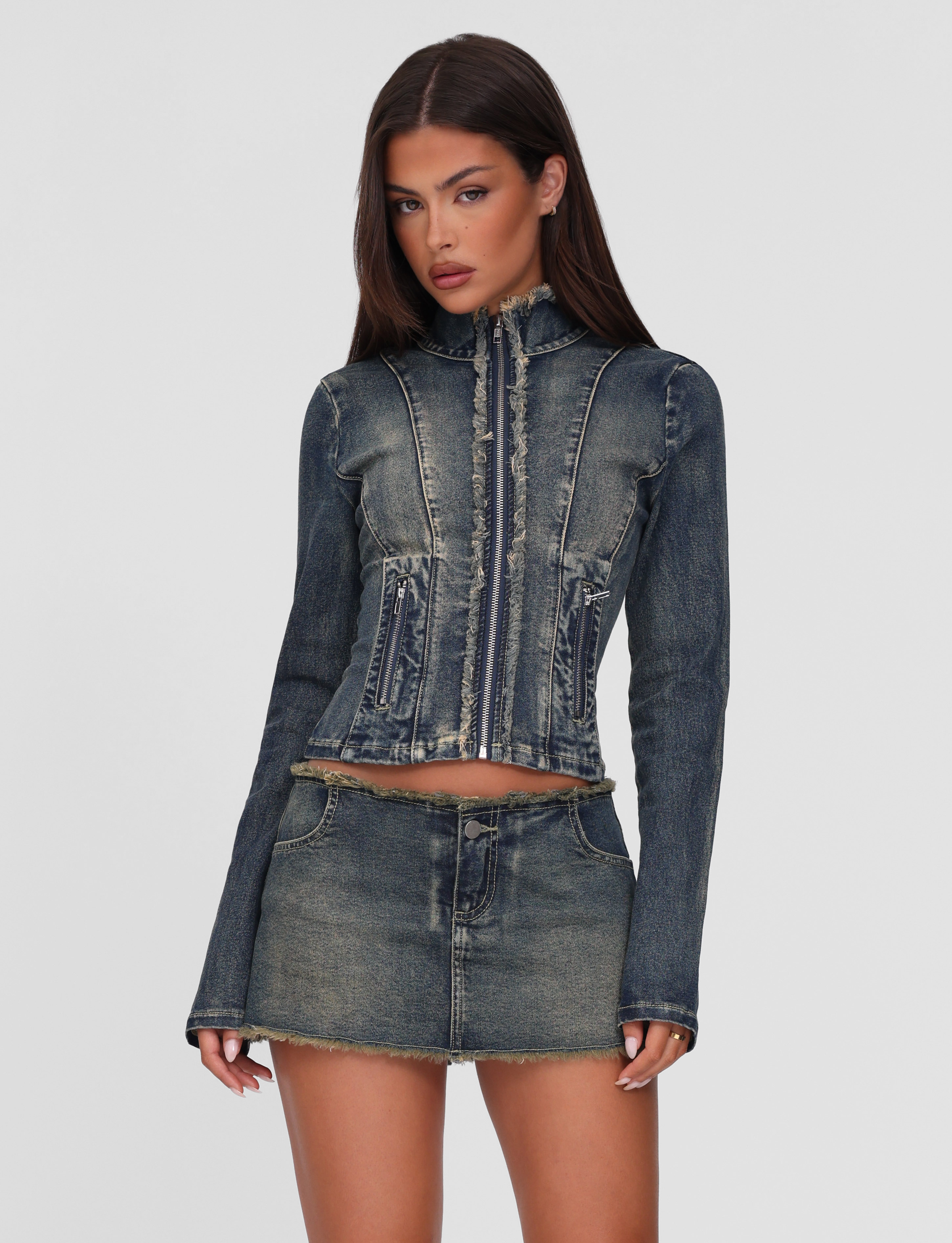 A woman with long brunette hair wears the TIGERMIST OPHELIE MINI SKORT in denim blue with a matching dark-wash, relaxed-fit denim jacket, standing against a plain light gray background.