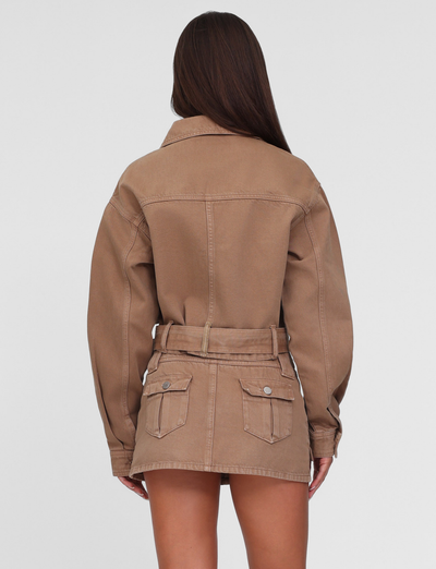 A woman with long brown hair faces away from the camera, wearing the TIGERMIST ZELIE JACKET in neutral sandblast washed tan, paired with a matching miniskirt. The background is plain white.