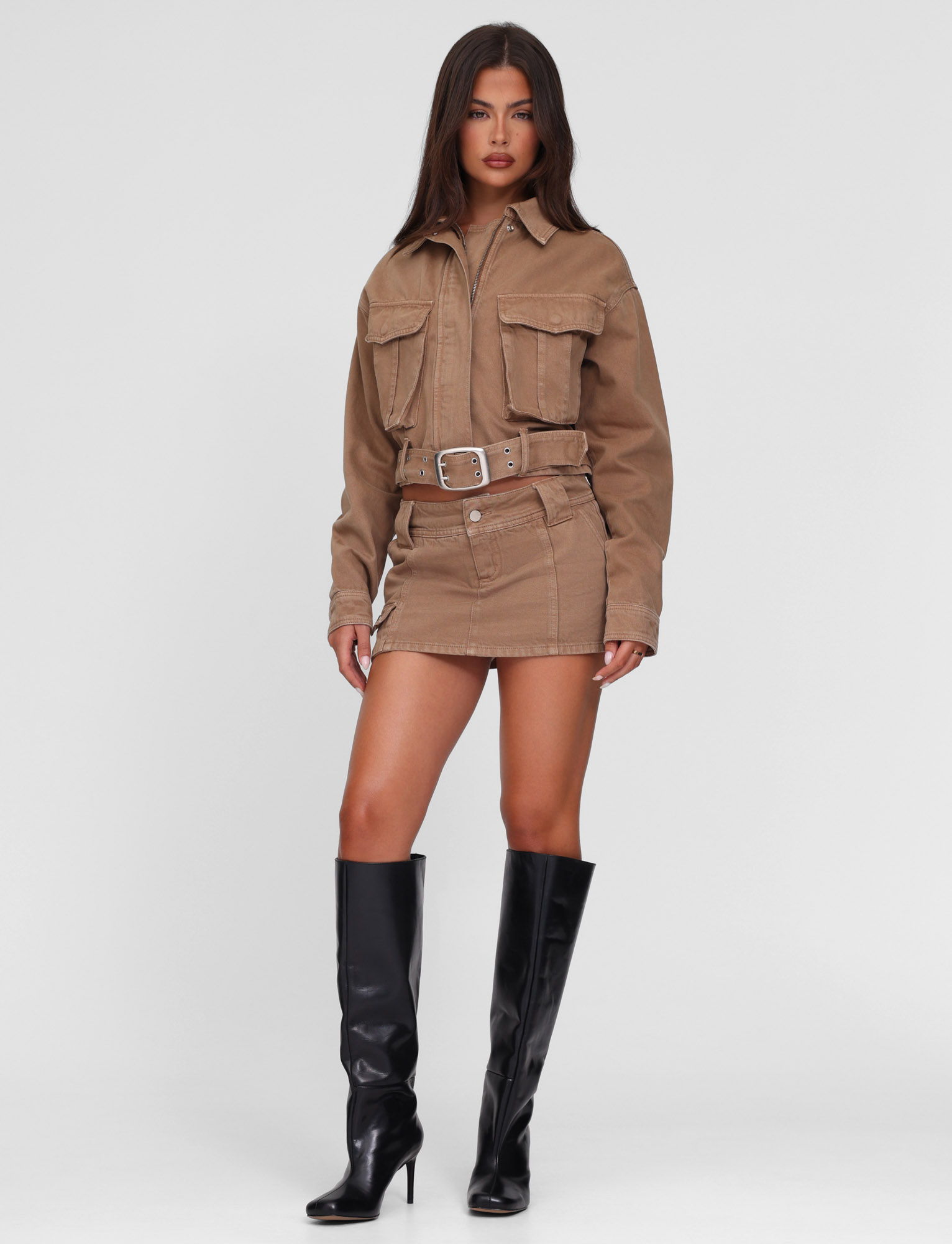 A woman with long dark hair and a neutral expression wears the TIGERMIST ZELIE JACKET in washed tan sandblast, styled with a matching tan mini skirt and black knee-high heeled boots, standing against a plain white background.