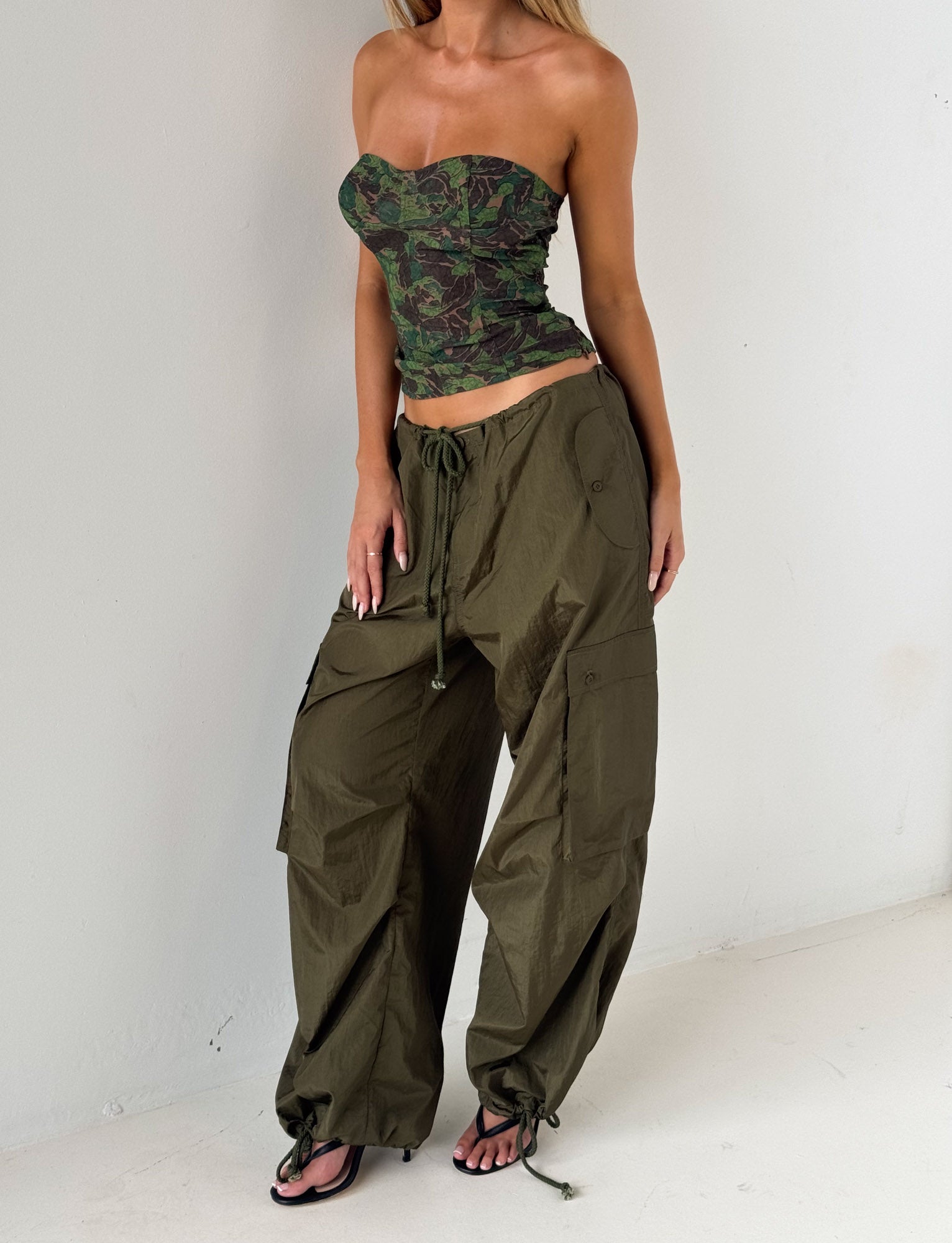 A woman stands against a white wall wearing the TIGERMIST VIOLETTE PANT in khaki green, styled with a green camo strapless top and black open-toe heels.