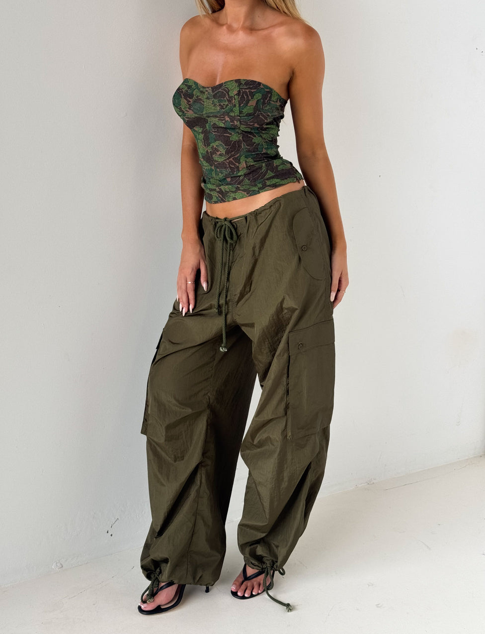 A woman stands against a white wall wearing the TIGERMIST VIOLETTE PANT in khaki green, styled with a green camo strapless top and black open-toe heels.