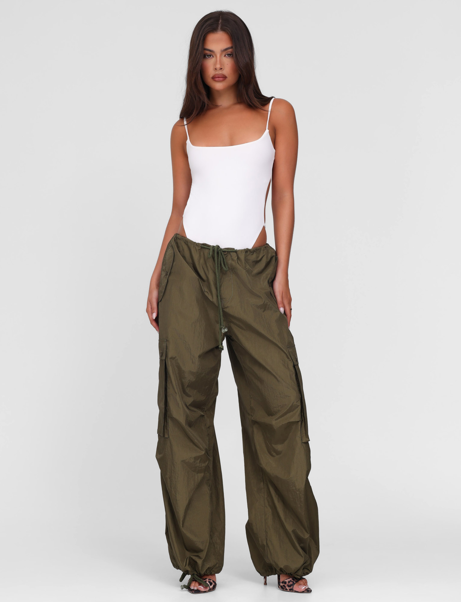 A woman with long dark hair wears the TIGERMIST VIOLETTE PANT in khaki green, styled with a white fitted tank bodysuit and black heeled sandals, standing against a plain background.