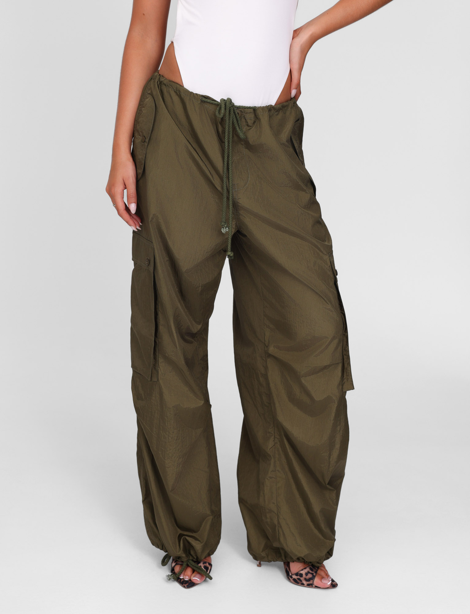 A person wears TIGERMIST's VIOLETTE PANT in khaki green, featuring a drawstring waist, side pockets, and adjustable hems, paired with a sleeveless white bodysuit and open-toe heels.