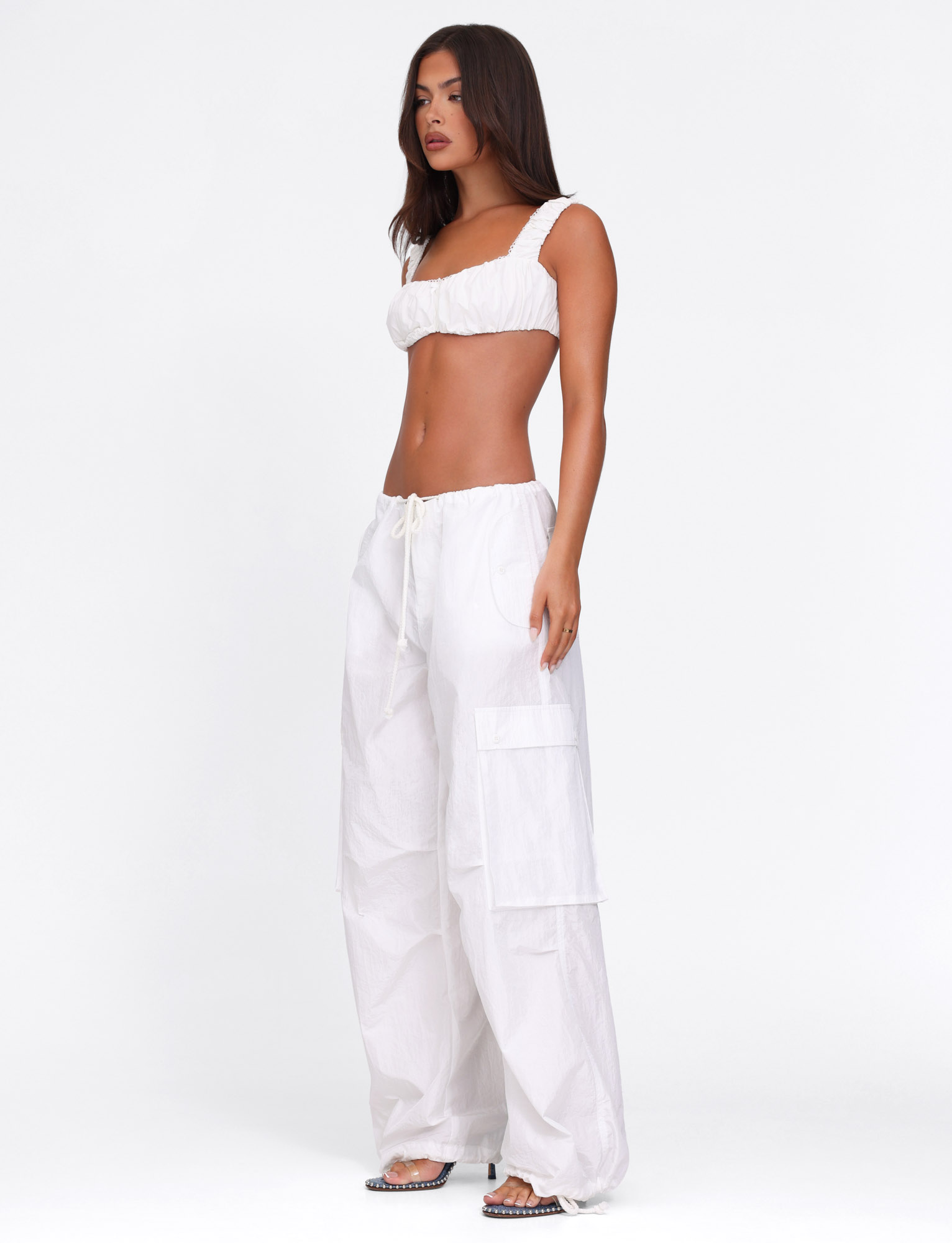 A woman with long, straight dark hair stands barefoot against a white background, wearing the TIGERMIST VIOLETTE PANT in white and a white cropped top with thick straps and an oversized fit.