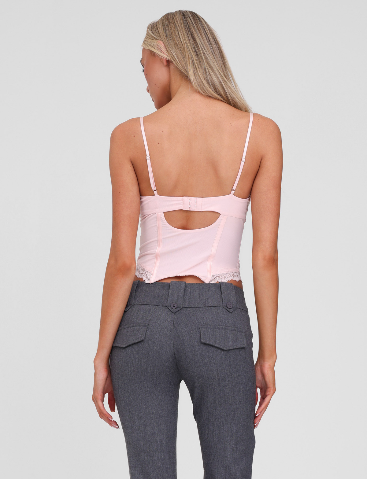 A woman with long blonde hair is shown from behind, wearing the TIGERMIST NILA CORSET TOP in soft pink with adjustable straps, paired with high-waisted gray pants featuring buttoned back pockets.