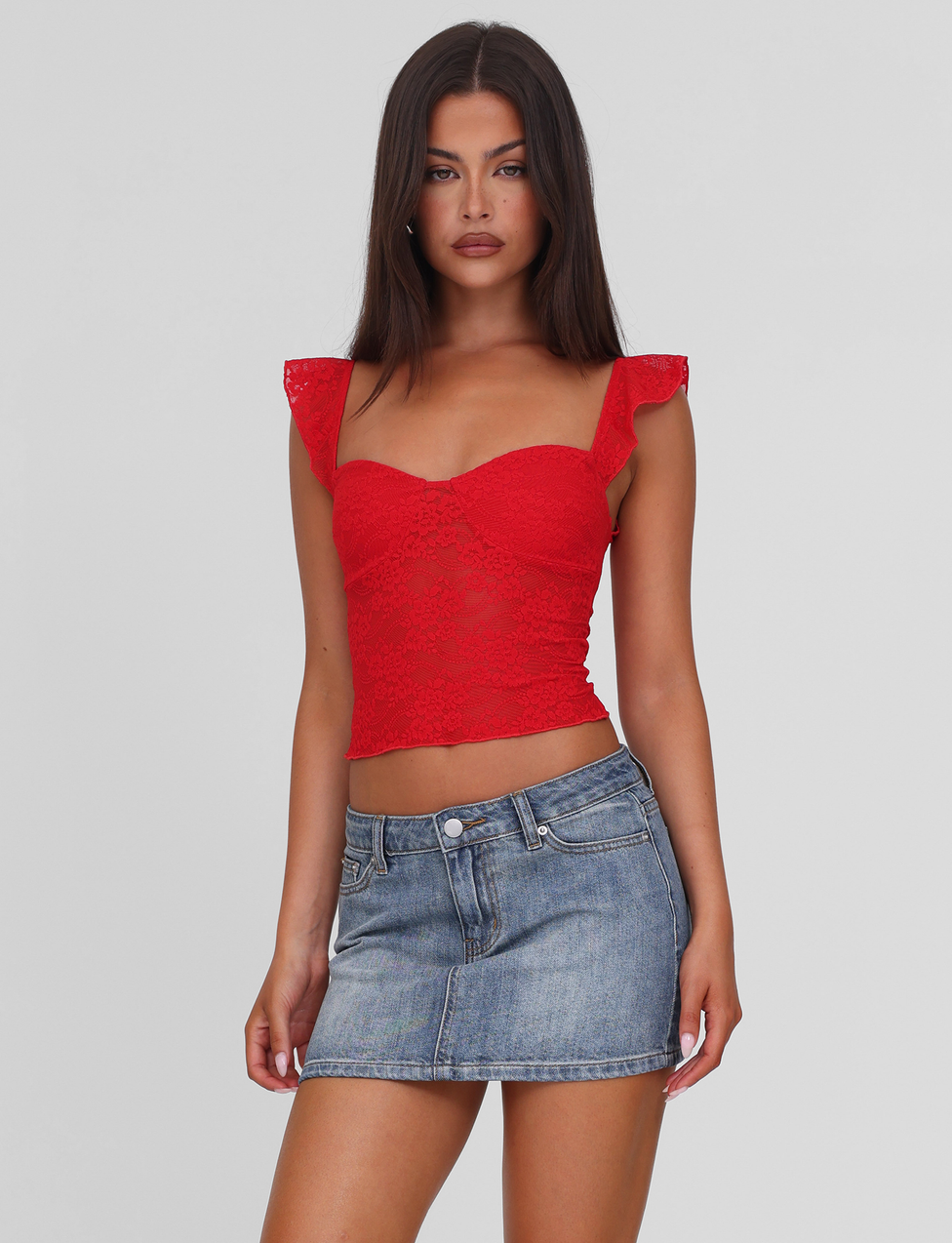 A woman with long dark hair wears the TIGERMIST LORNA TOP in red and a short blue denim skirt, standing against a plain light gray background and looking forward with a neutral expression.