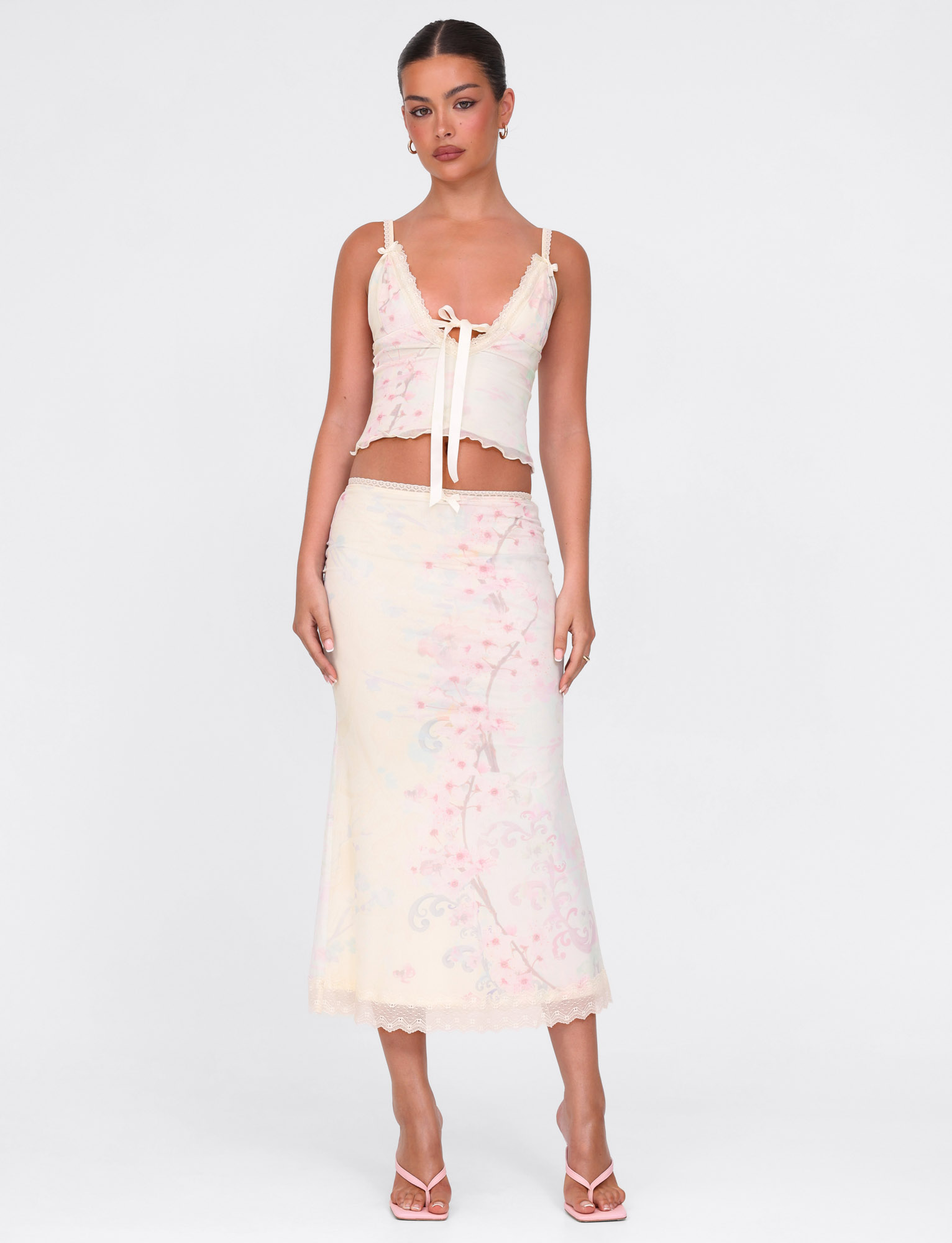A woman wears the TIGERMIST LONIE TOP in Neutral Floral Washed Cherry Blossom, a sleeveless cropped top with lace trim and bow detail, paired with a coordinating pastel high-waisted skirt and open-toe heels, standing against a plain background.