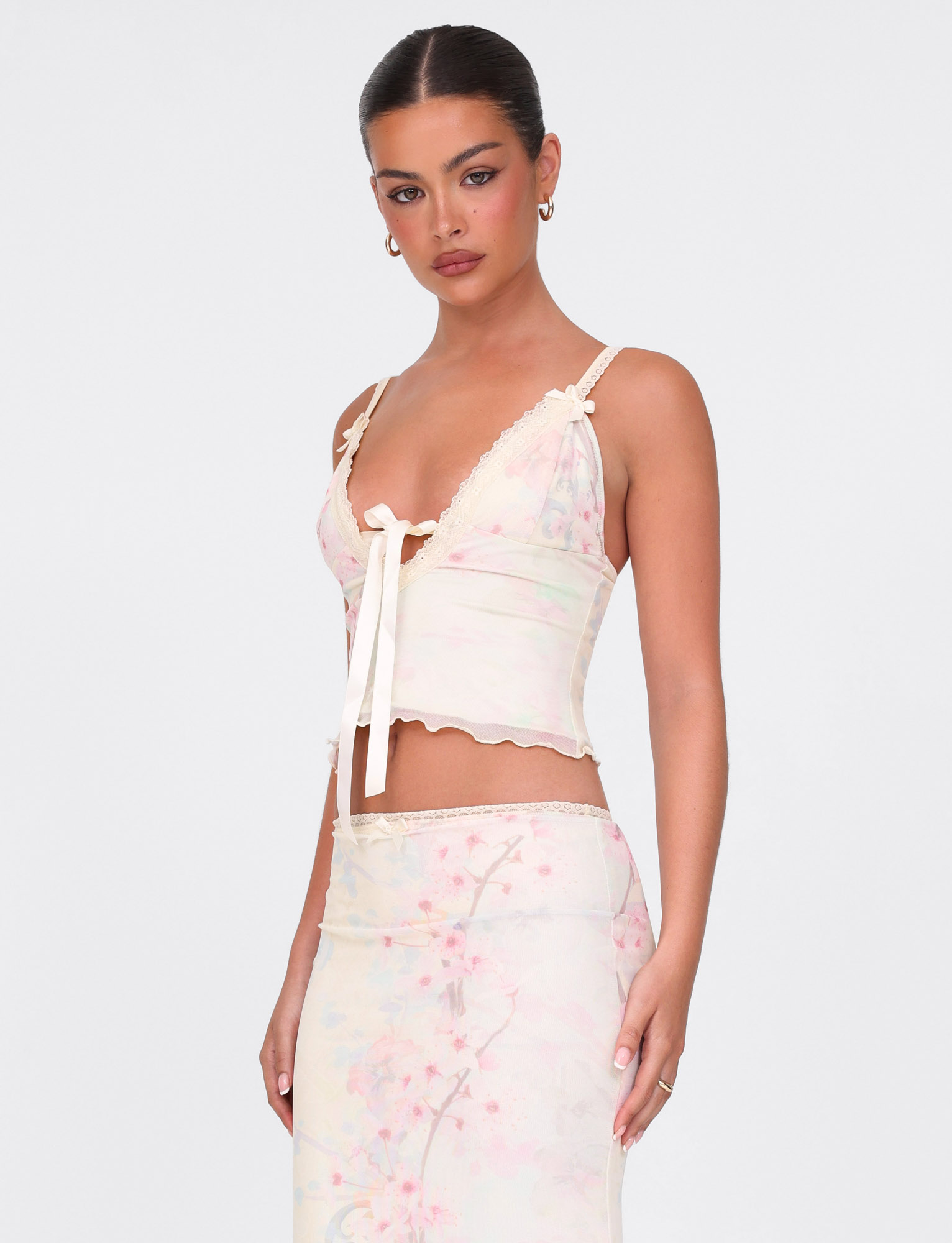 A woman with slicked-back hair wears the TIGERMIST LONIE TOP in Neutral Floral Washed Cherry Blossom, featuring lace trim and bow details, paired with a matching skirt, standing confidently against a plain white background.