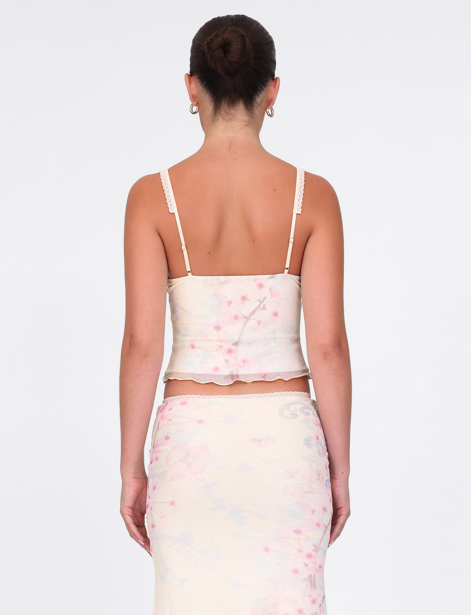 A woman with dark hair in a bun wears the TIGERMIST LONIE TOP in neutral washed cherry blossom floral, paired with a matching skirt and gold hoop earrings, standing against a plain white background.