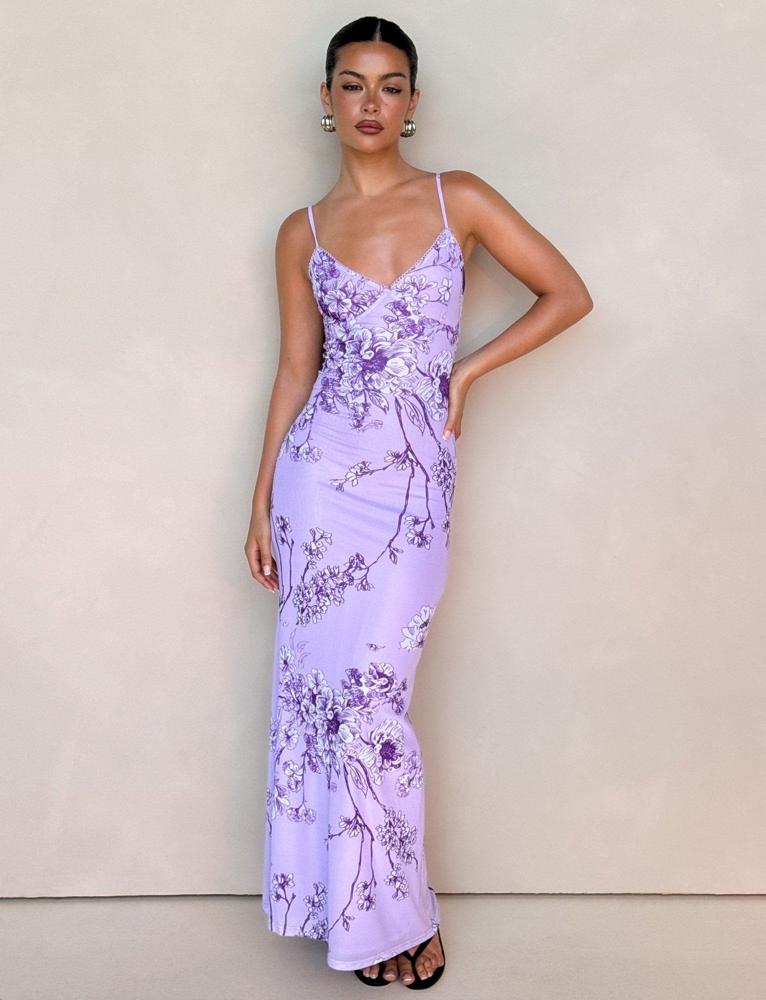 A person stands against a plain background wearing a TIGERMIST Julianna Dress in purple ceramic floral, a form-fitting gown with thin straps. They have their hand on their hip, wear hoop earrings and black open-toe heels, and have their hair neatly tied back.
