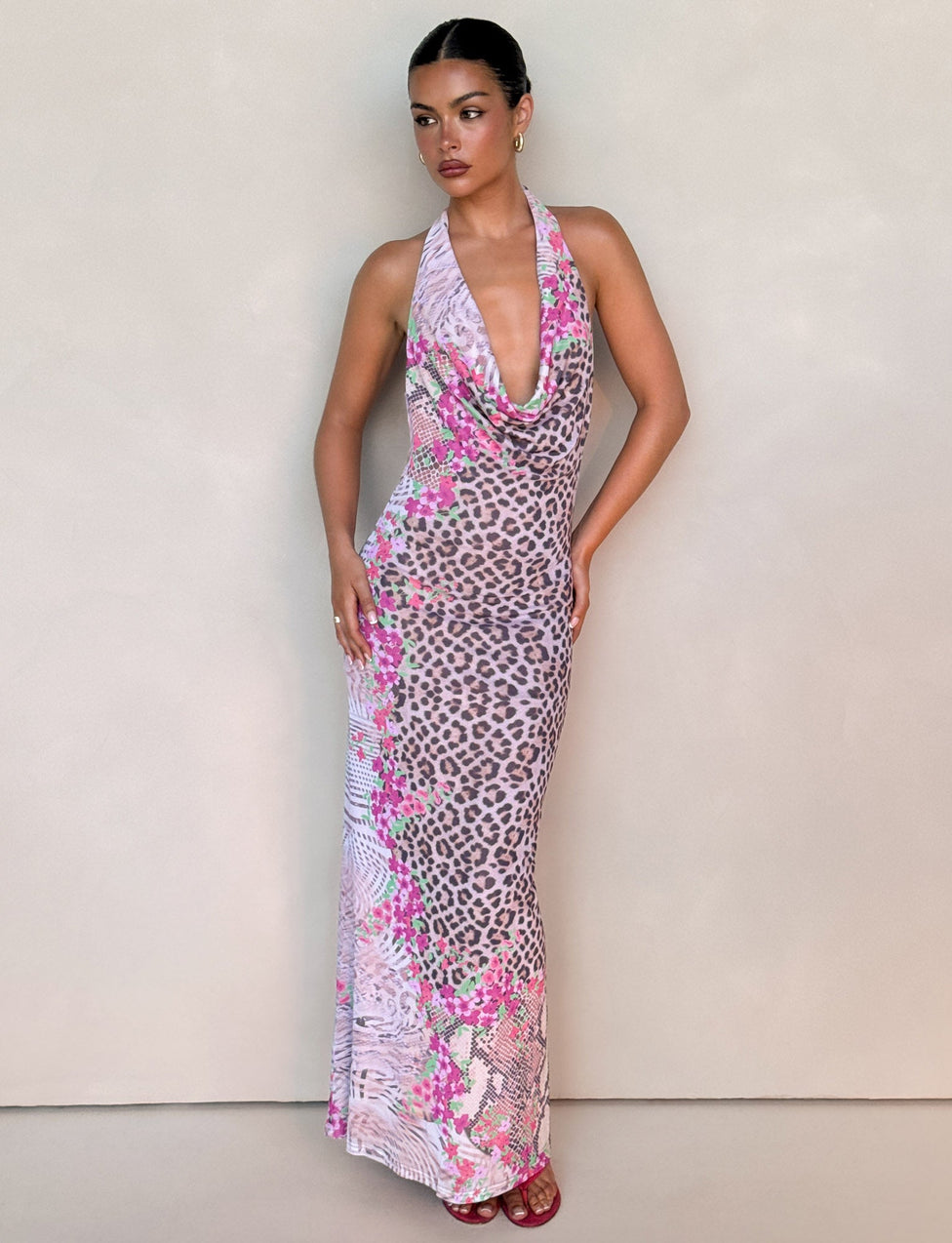 A woman stands against a beige wall, wearing the TIGERMIST CATALINA MAXI DRESS in Neutral Animal Floral Border. The sleeveless, halter-neck, backless dress features a pink and gray animal print. Her hair is pulled back and she wears pink shoes.