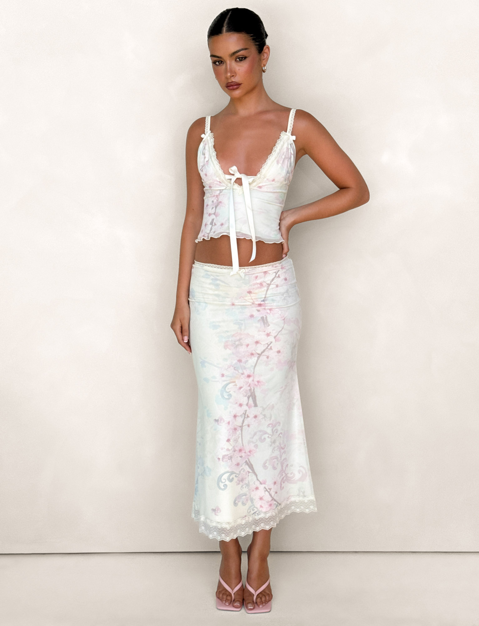A woman stands against a light background wearing the TIGERMIST LONIE MIDI SKIRT in Washed Cherry Blossom, a pastel floral two-piece with lace-trim top and exclusive printed mesh skirt. She has dark hair pulled back and pink strappy heels.