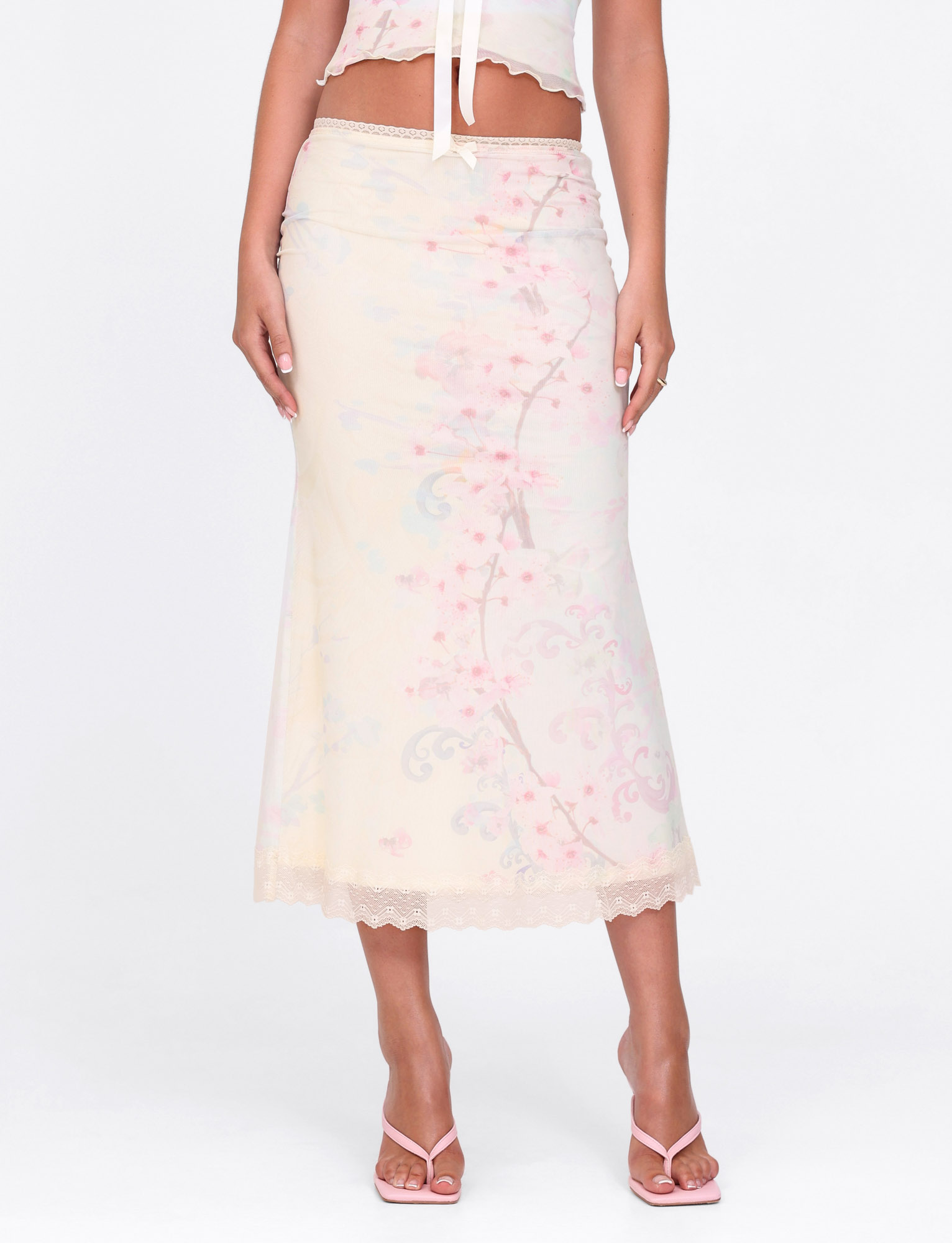 A person wears the TIGERMIST LONIE MIDI SKIRT in Washed Cherry Blossom, featuring a light floral print and lace trim, styled with a matching crop top and strappy sandals for a soft, feminine pastel look.