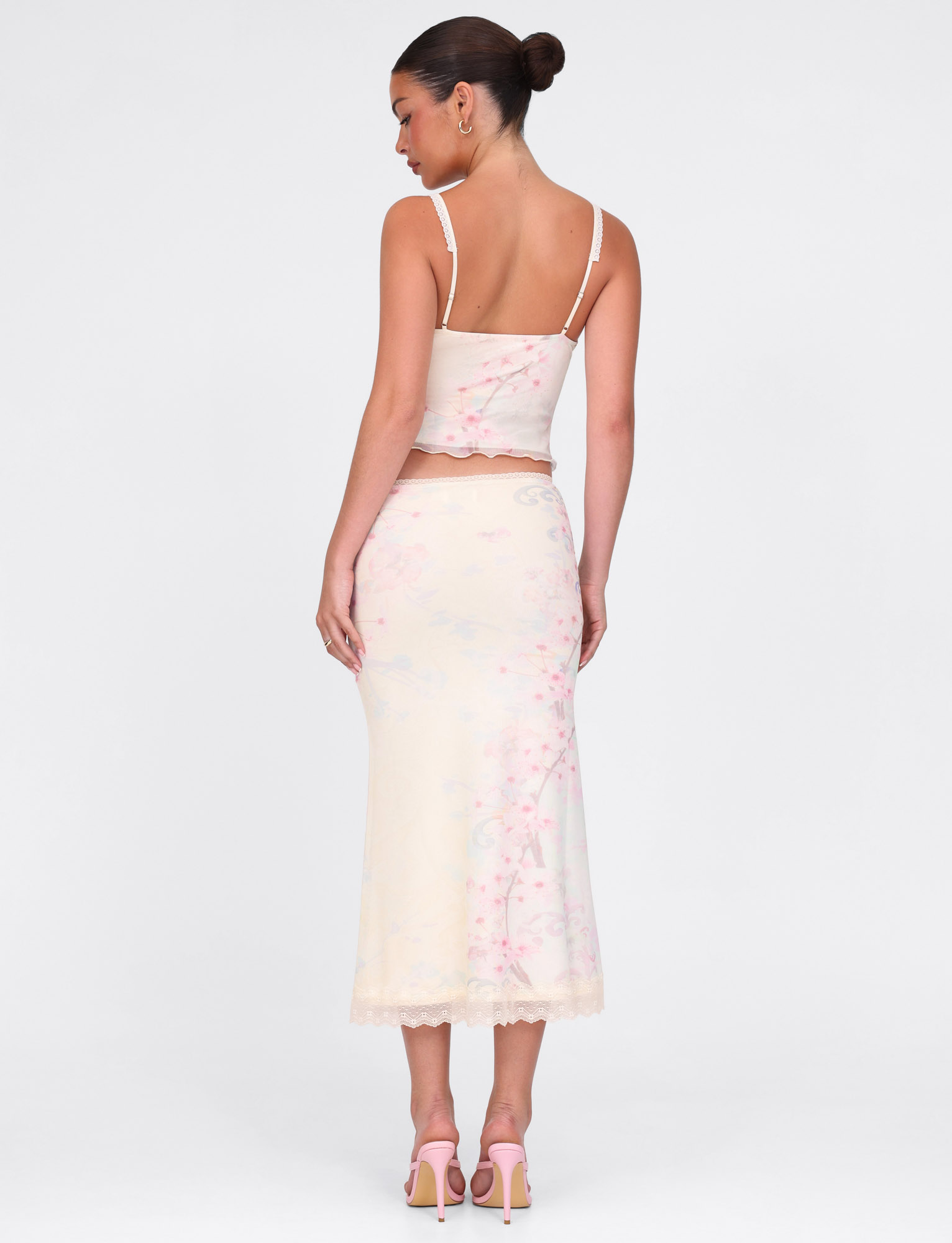 A woman faces away in a cream spaghetti strap top and the TIGERMIST LONIE MIDI SKIRT—Washed Cherry Blossom floral print with lace trim—paired with pink heels. Her hair is styled in a neat bun.
