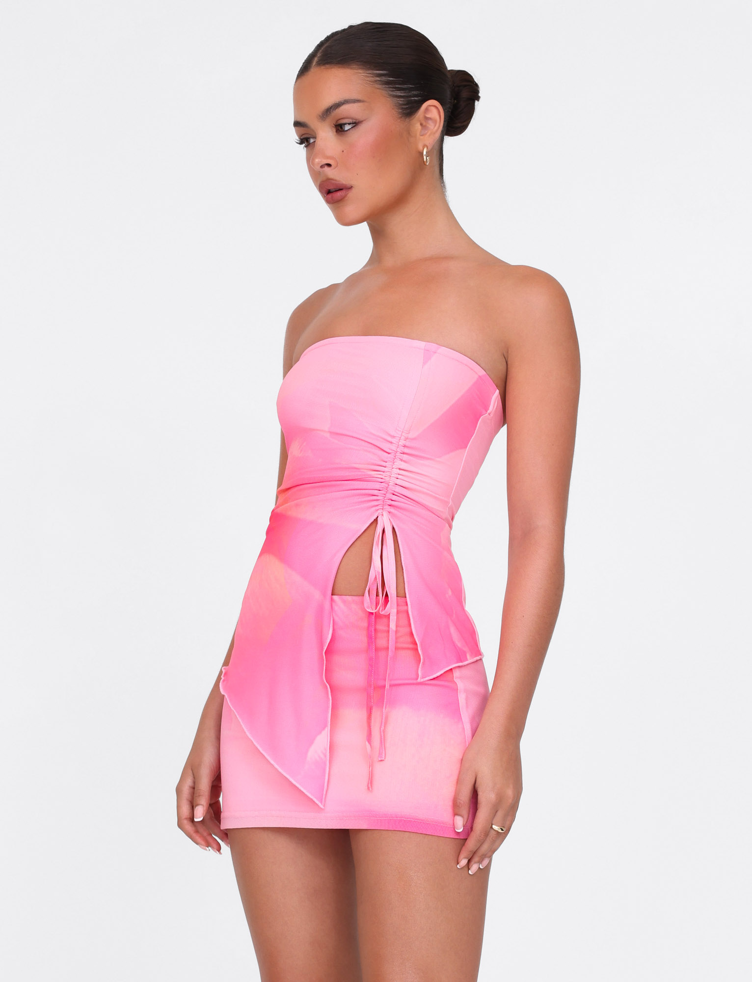 A woman stands against a plain white background wearing the TIGERMIST SARIAH MINI SKIRT in pink blossom macro floral, featuring a layered skirt, side cutout, ruched details, and high stretch fabric.