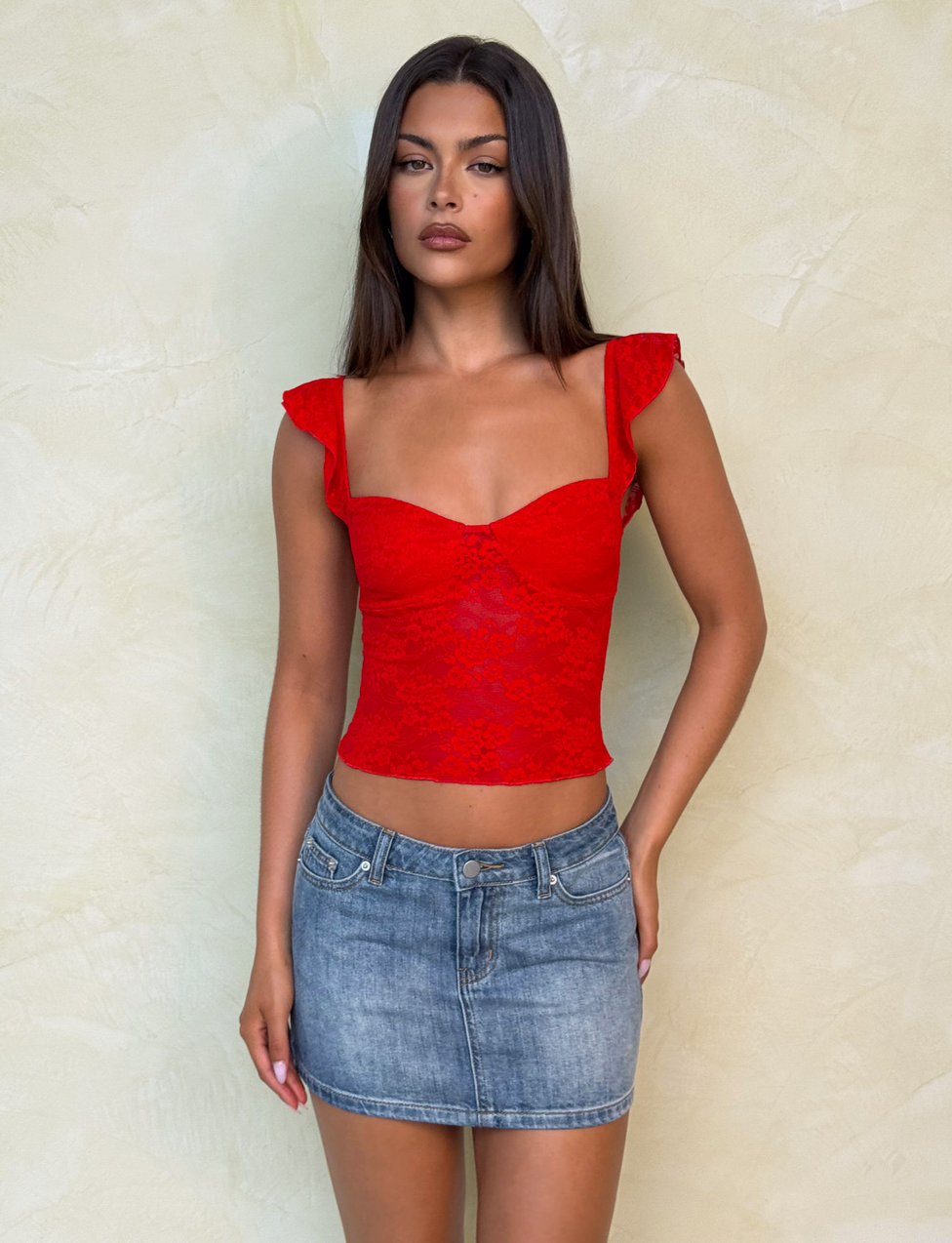 A woman with long, straight dark hair and a neutral expression stands against a light textured wall, wearing TIGERMIST's MICHAYLA MINI SKIRT in mid wash blue denim and a fitted red lace top with ruffled sleeves.