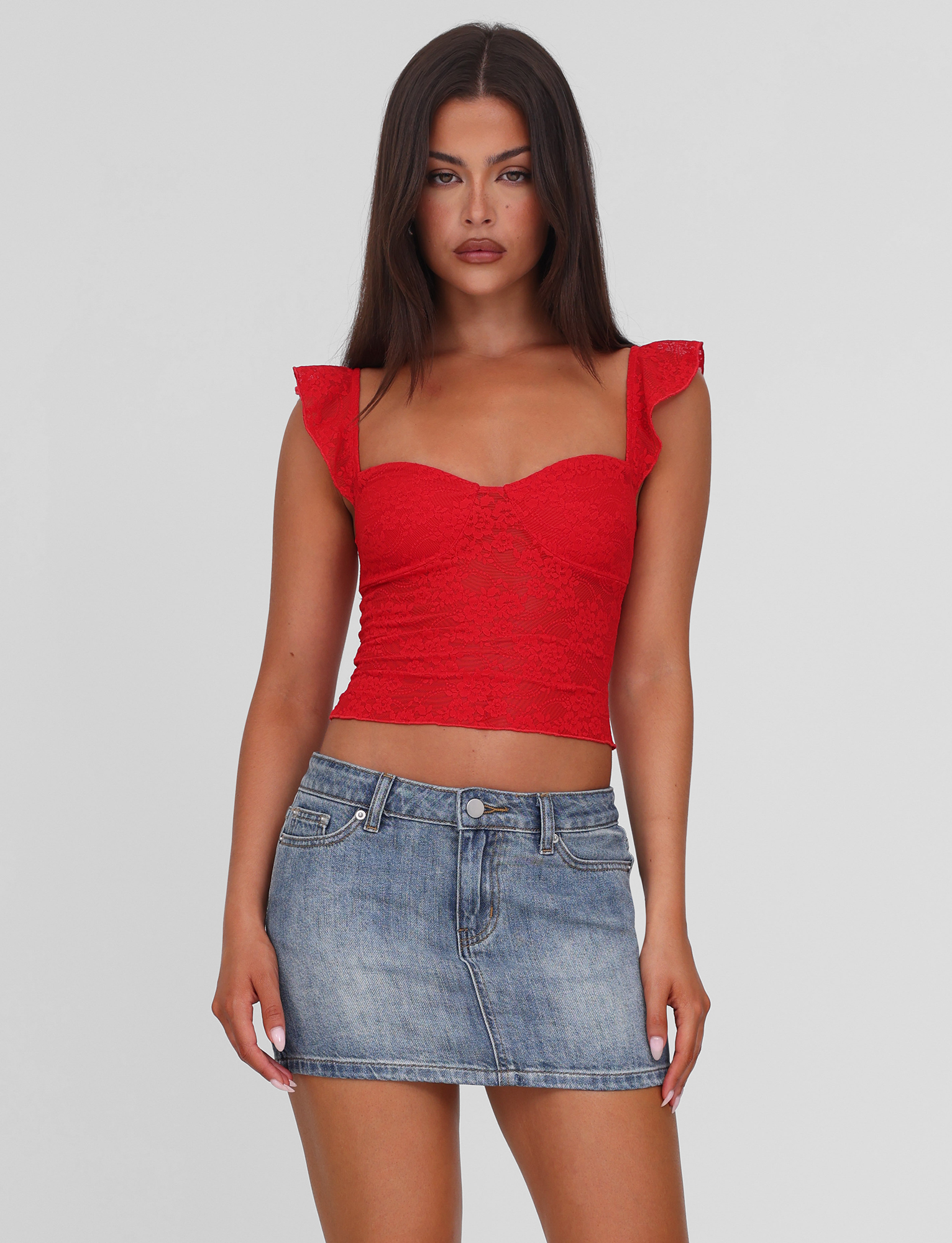 A woman with long brown hair wears a red lace crop top and the TIGERMIST MICHAYLA MINI SKIRT in mid wash, styled with five pockets, standing against a plain white background.