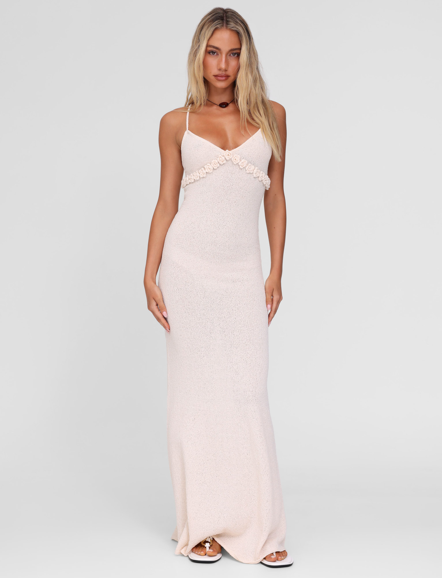 A woman with long blonde hair models the TIGERMIST LAVELLE MAXI DRESS in cream, a light pink, sleeveless, floor-length gown with floral detail and adjustable crisscross straps, standing against a plain white background.