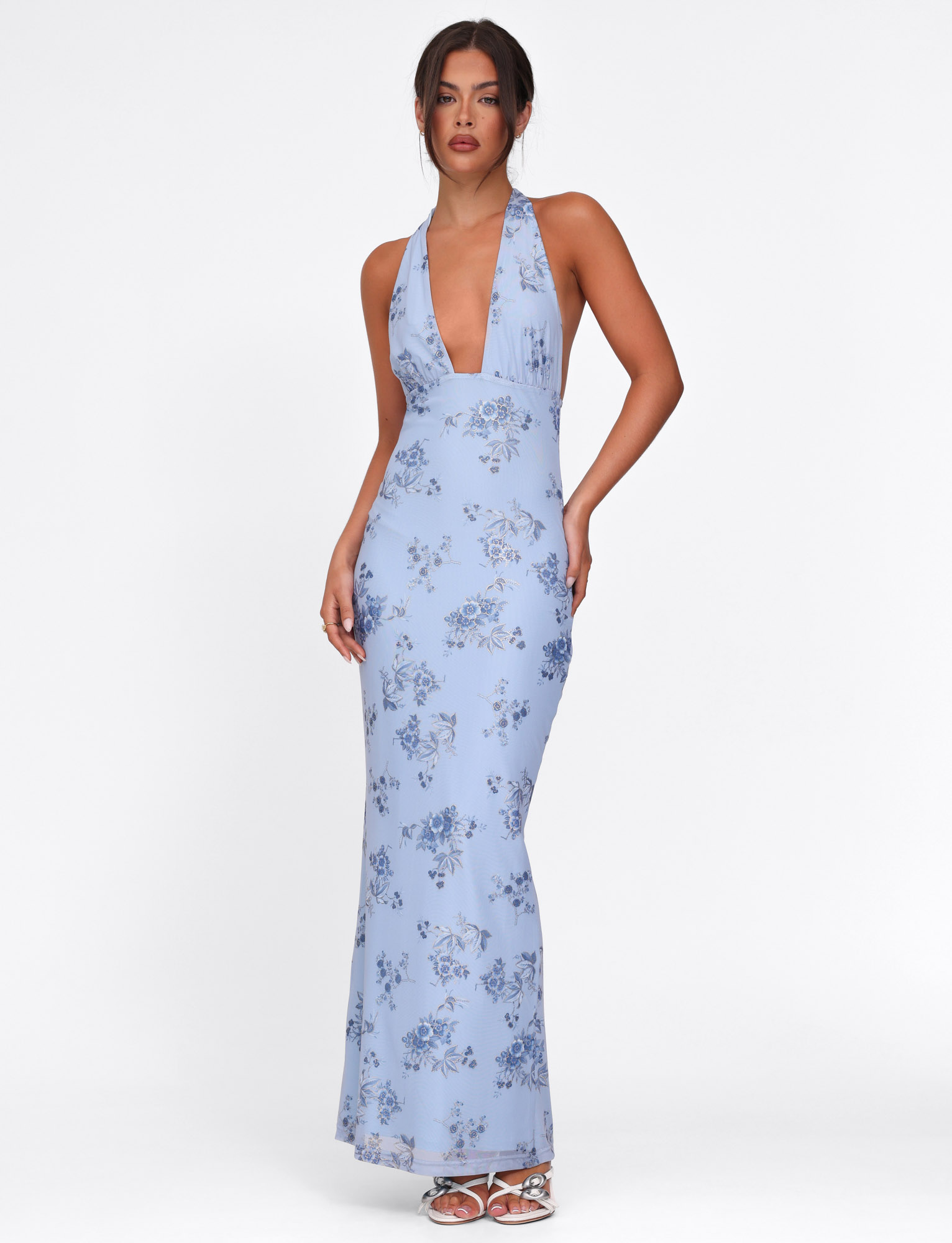 A woman stands against a white background wearing the TIGERMIST PHAEDRA MAXI DRESS in blue floral, featuring a halter neck, deep V-neckline, and low open back.