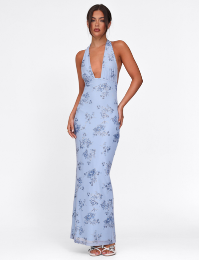 A woman stands against a white background wearing the TIGERMIST PHAEDRA MAXI DRESS in blue floral, featuring a halter neck, deep V-neckline, and low open back.