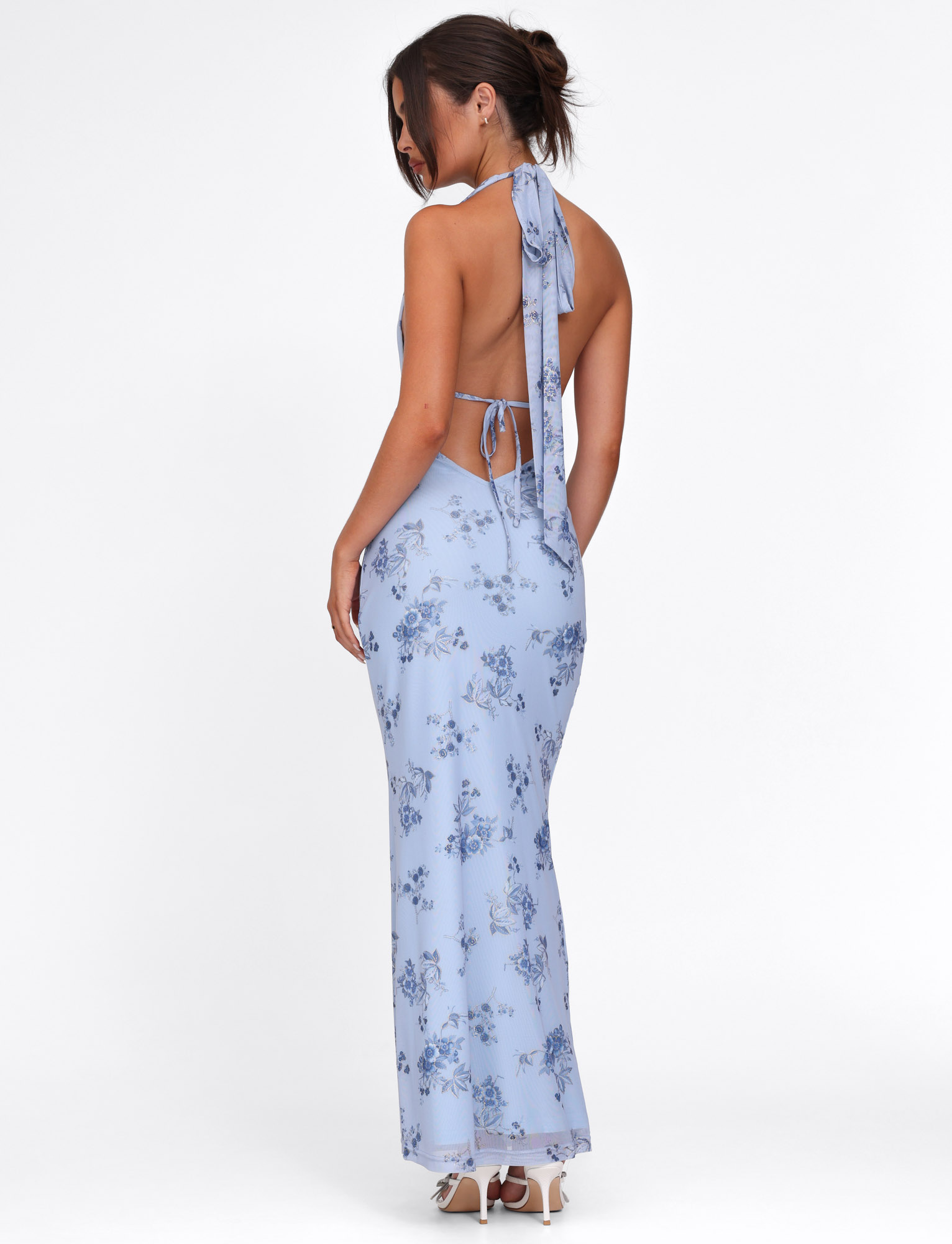A woman stands sideways, looking down, wearing the TIGERMIST PHAEDRA MAXI DRESS in blue floral with a halter neck and low open back, her hair in a loose bun and paired with white high-heeled shoes.