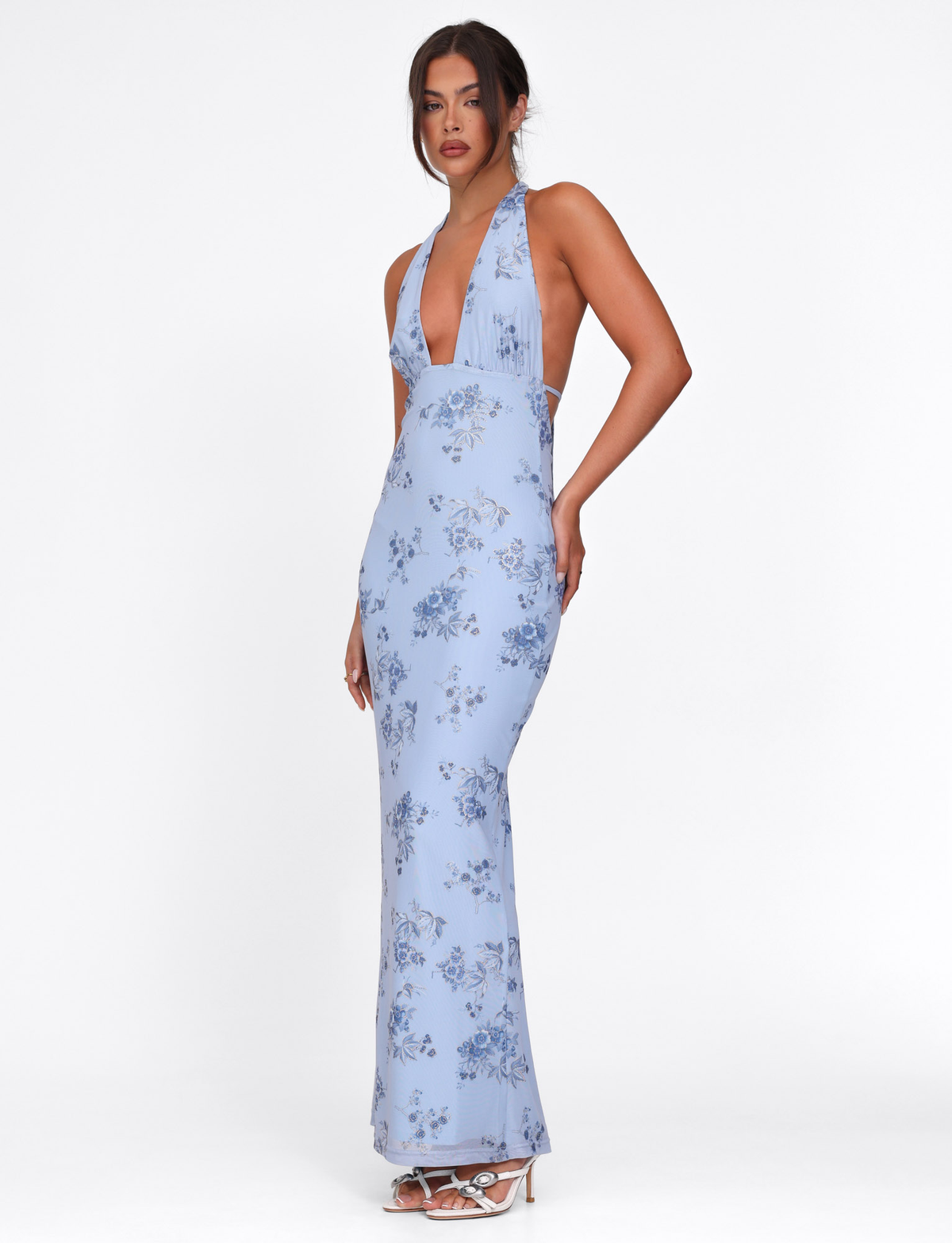 A woman models the TIGERMIST PHAEDRA MAXI DRESS in blue floral, featuring a halter neck, deep V neckline, and low open back. She pairs it with white heeled sandals against a plain white background.