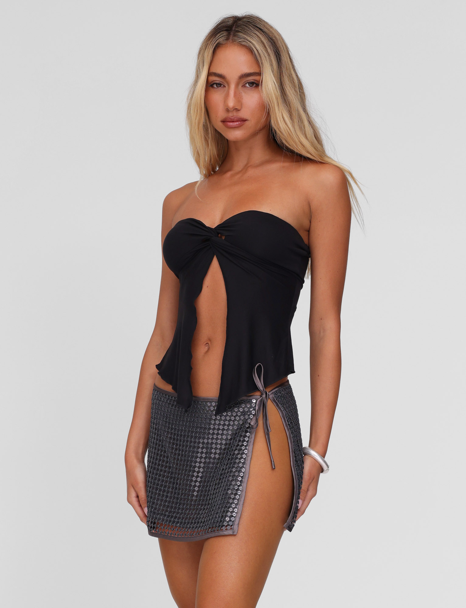 A woman with long blonde hair wears a strapless black top and the TIGERMIST VERENA MINI SKIRT in steel grey, featuring an asymmetrical hem and low-rise waist, posing against a plain light background.