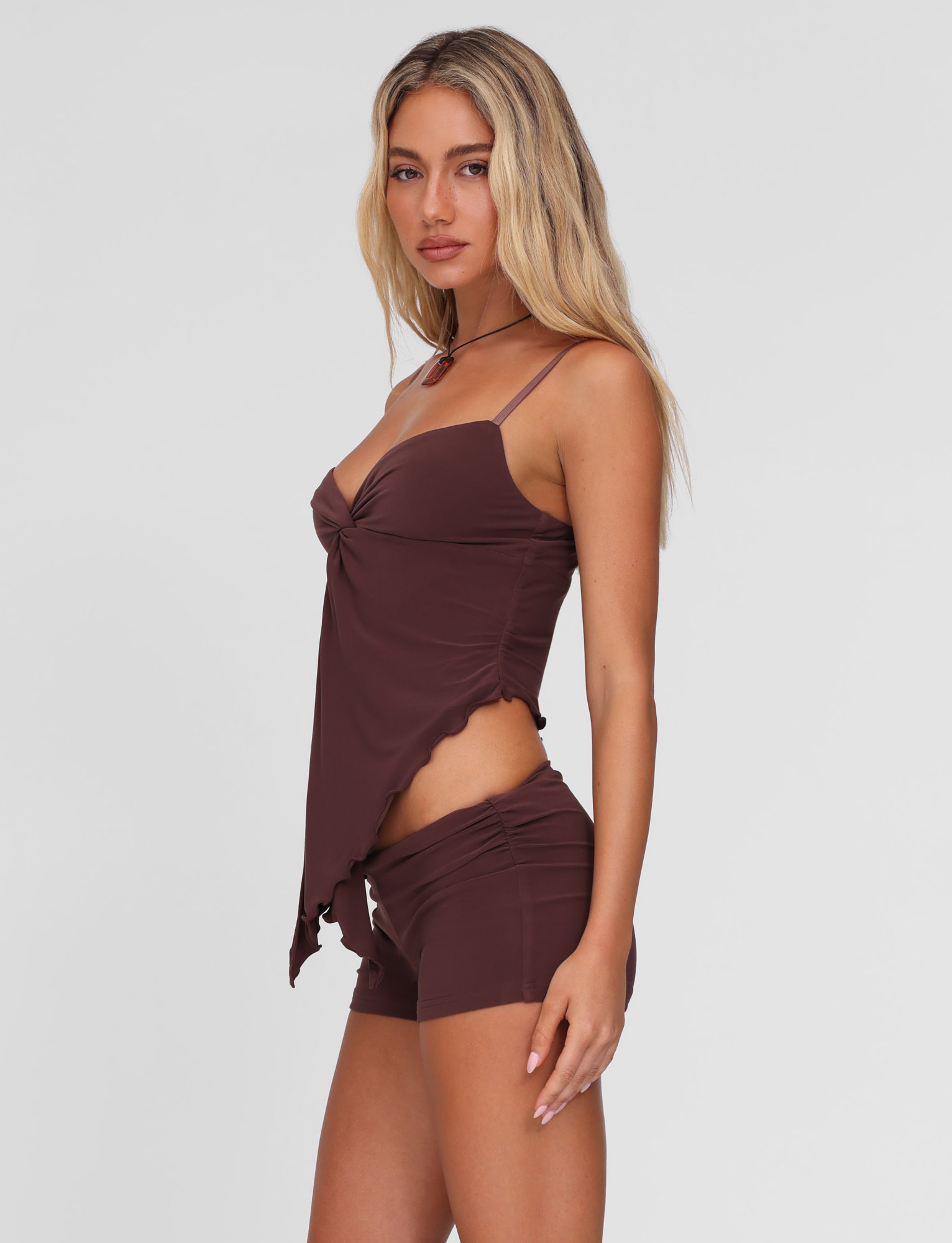 A woman with long blonde hair models the TIGERMIST JOVIENNE TOP - BROWN : CHOCOLATE, a chocolate camisole with thin straps, twist front detail, and asymmetric hem, styled with matching brown shorts against a light gray background.