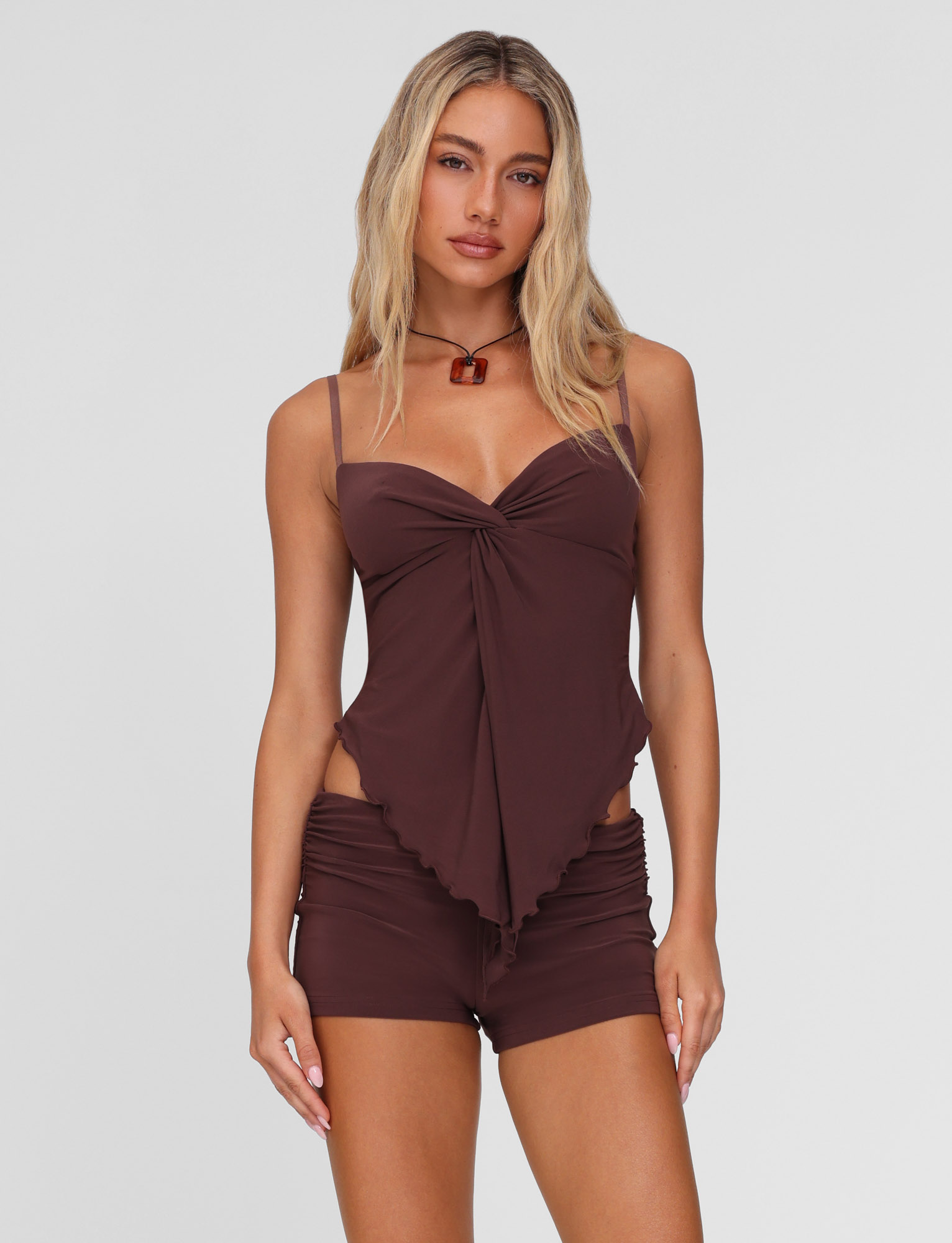 A woman with long blonde hair models the TIGERMIST JOVIENNE SHORT - BROWN: CHOCOLATE, featuring an asymmetrical, sleeveless spaghetti strap top and matching micro mini shorts, posing against a plain white background.