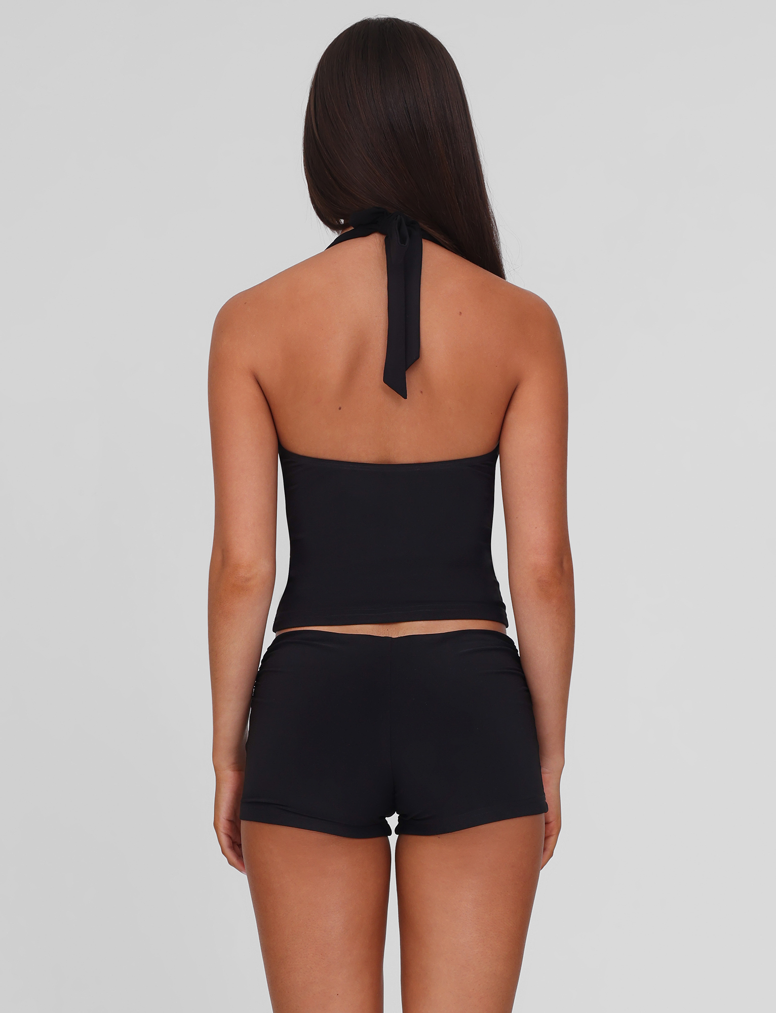 A woman with long brown hair stands with her back to the camera, wearing a black halter top and TIGERMIST JOVIENNE SHORT - BLACK, which are fitted, mid-rise jersey mini shorts, set against a plain light gray background.