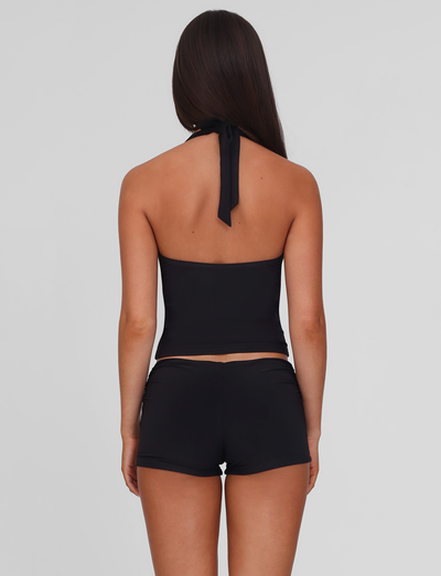 A woman with long brown hair stands with her back to the camera, wearing a black halter top and TIGERMIST JOVIENNE SHORT - BLACK, which are fitted, mid-rise jersey mini shorts, set against a plain light gray background.