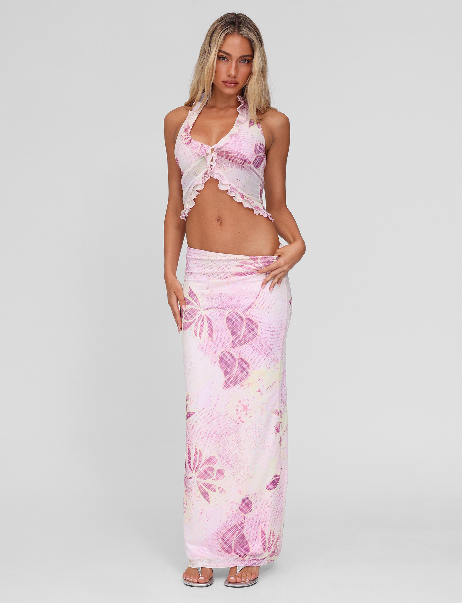 A woman with long blonde hair wears the TIGERMIST DALILA MIDI SKIRT in Grunge Floral Magenta with a matching pink and white floral halter crop top against a plain background, paired with sandals.