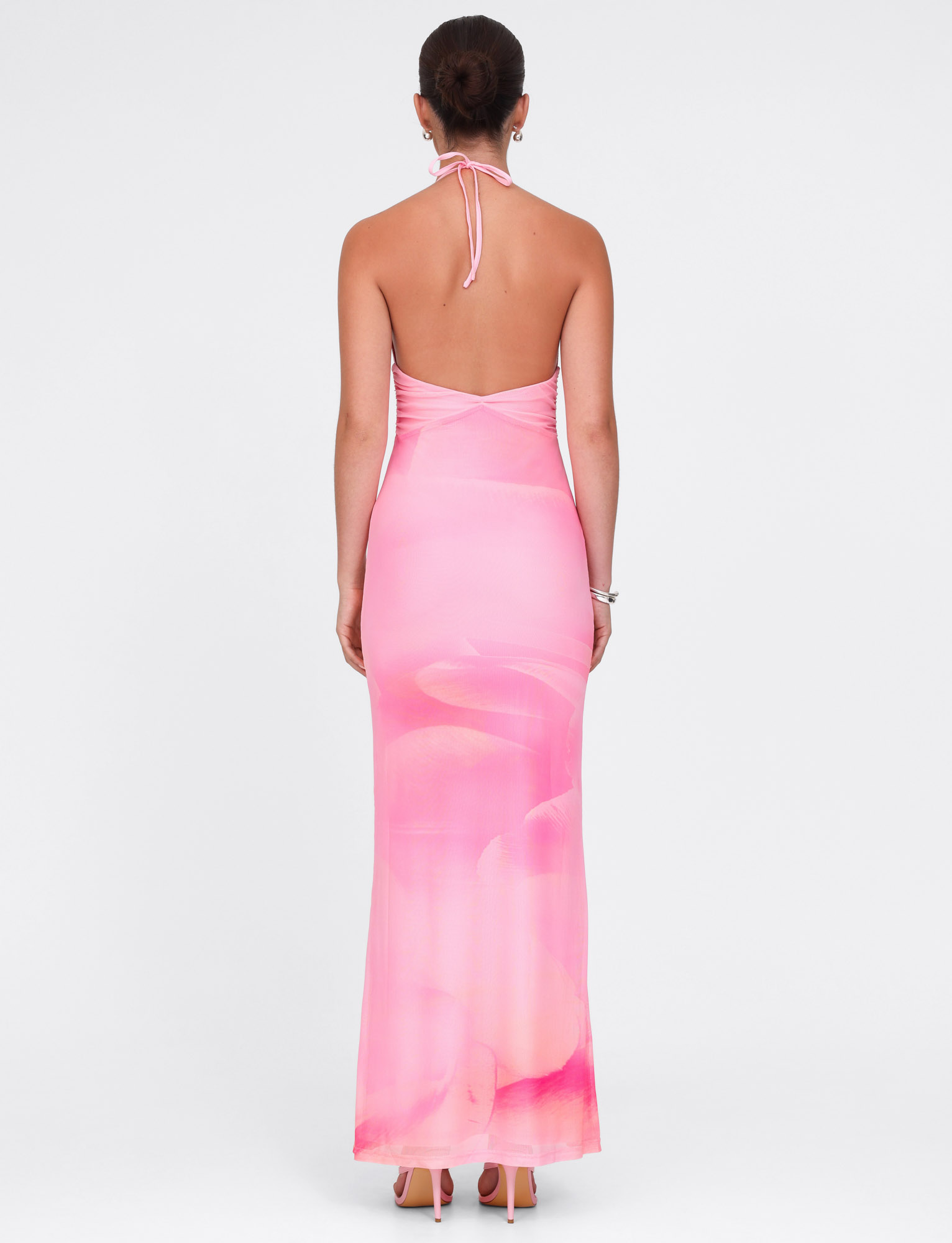 A woman faces away, wearing the TIGERMIST DENAE MAXI DRESS in Pink Blossom floral. Her hair is in a neat bun, styled with high heels and a bracelet. The background is plain white.