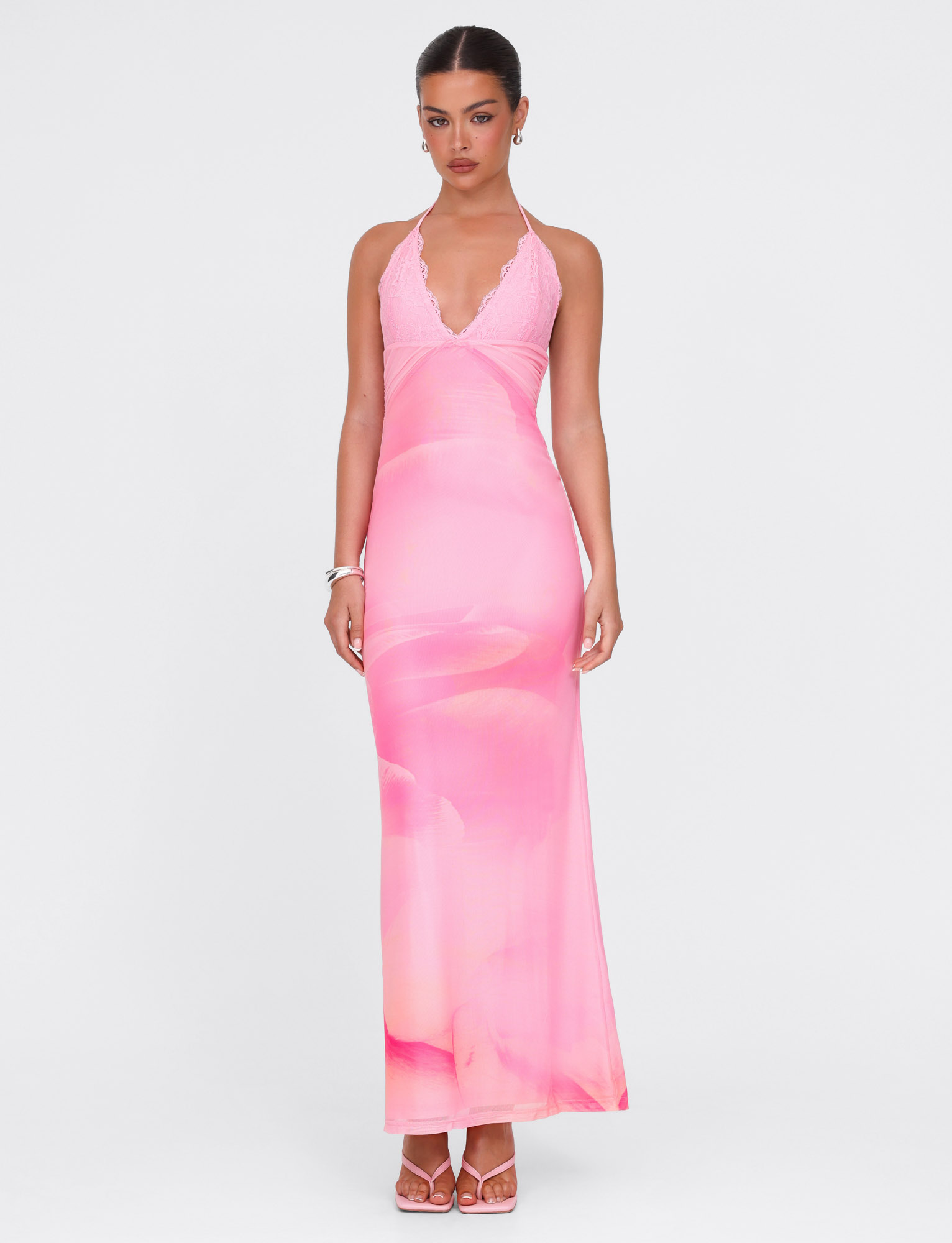 A woman stands against a white background wearing the TIGERMIST DENAE MAXI DRESS in Pink Blossom with matching heels and silver bracelets. Her hair is tied back as she looks at the camera, radiating effortless elegance.