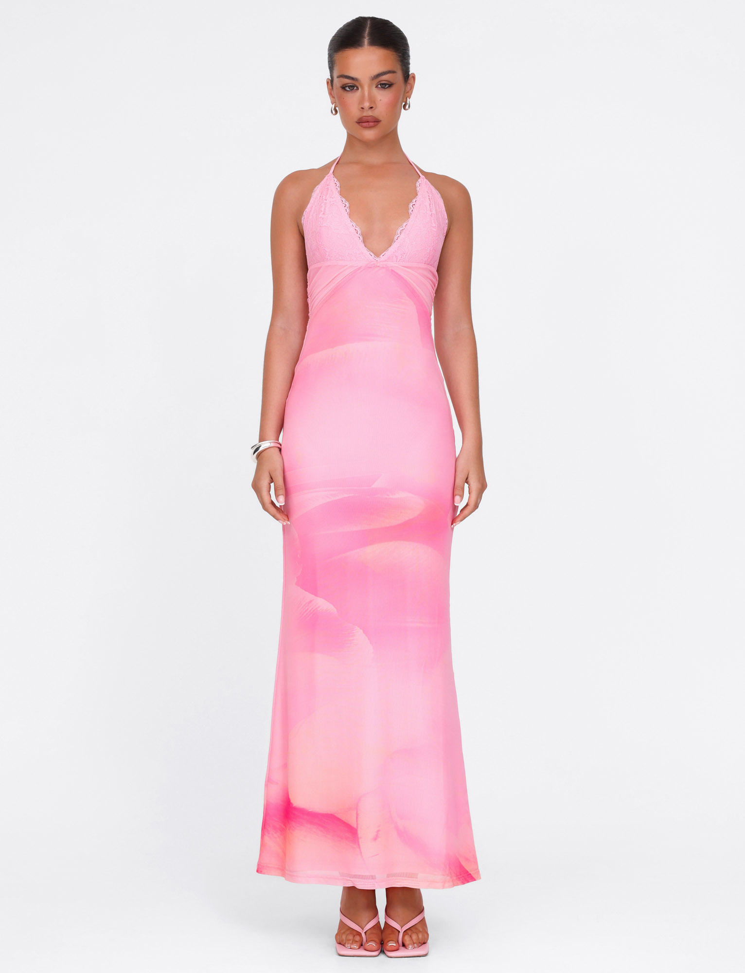 A woman wears the TIGERMIST DENAE MAXI DRESS in Pink Blossom, featuring a fitted silhouette, halter neckline, deep V cut, and subtle floral lace pattern. She styles it with pink sandals, simple jewelry, and a pulled-back hairstyle against a white background.