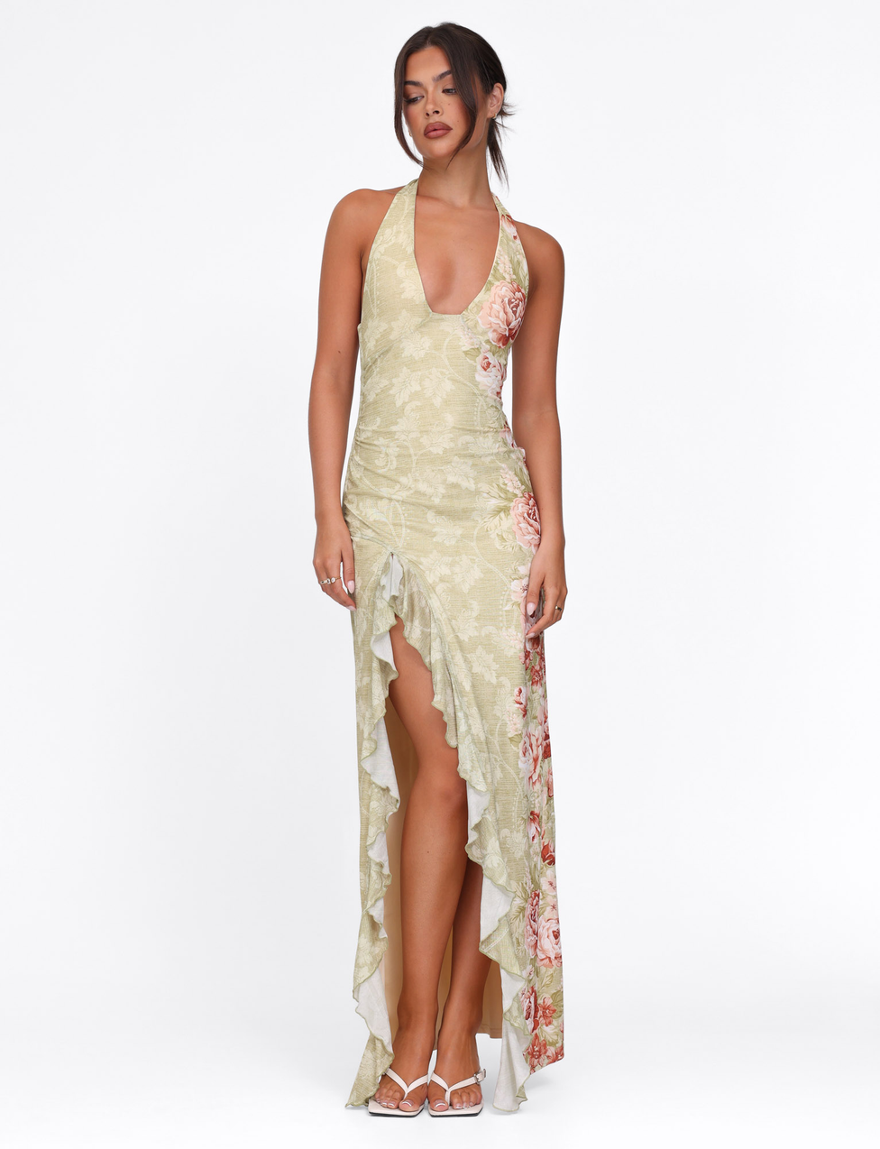 A woman poses against a white background in the TIGERMIST SIRENA MAXI DRESS in yellow floral rose stripe, featuring a halter neck, deep neckline, high split, ruffled details, and white strappy heels. Her hair is styled in a loose updo.