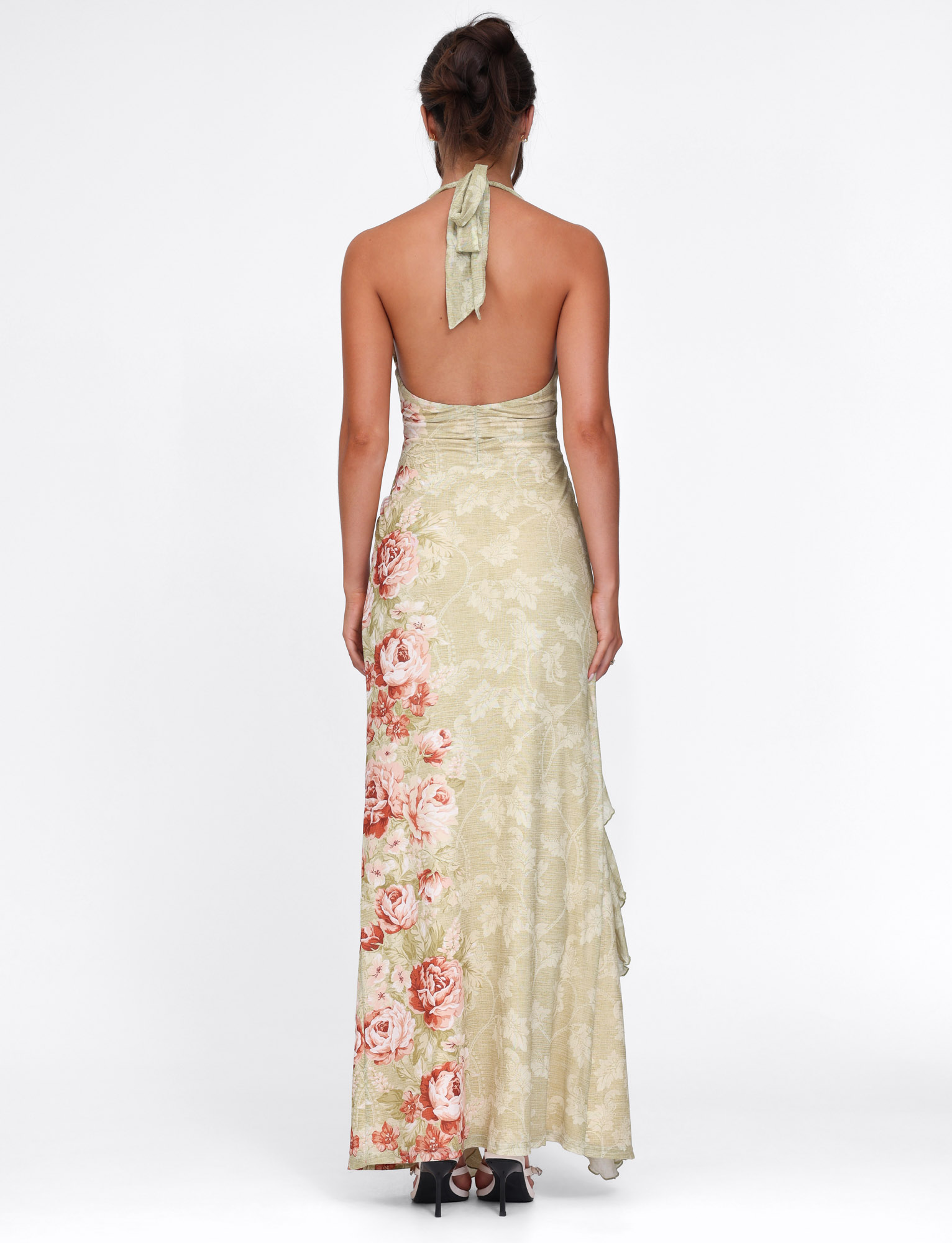 A woman with brown hair up wears the TIGERMIST SIRENA MAXI DRESS in yellow with a rose stripe floral print, featuring a halter neck and backless design. She pairs it with black heels, standing against a white background.