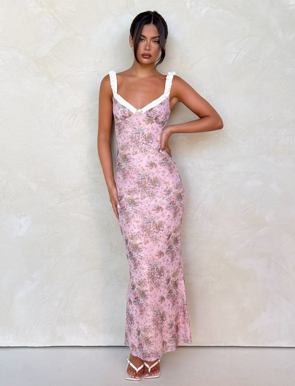 A woman stands against a textured light wall wearing the TIGERMIST AMARAN DRESS - PINK : CAMO FLORAL, a bodycon maxi with white ruffled straps. Her dark hair is in a loose updo and she pairs the dress with white strappy heels.