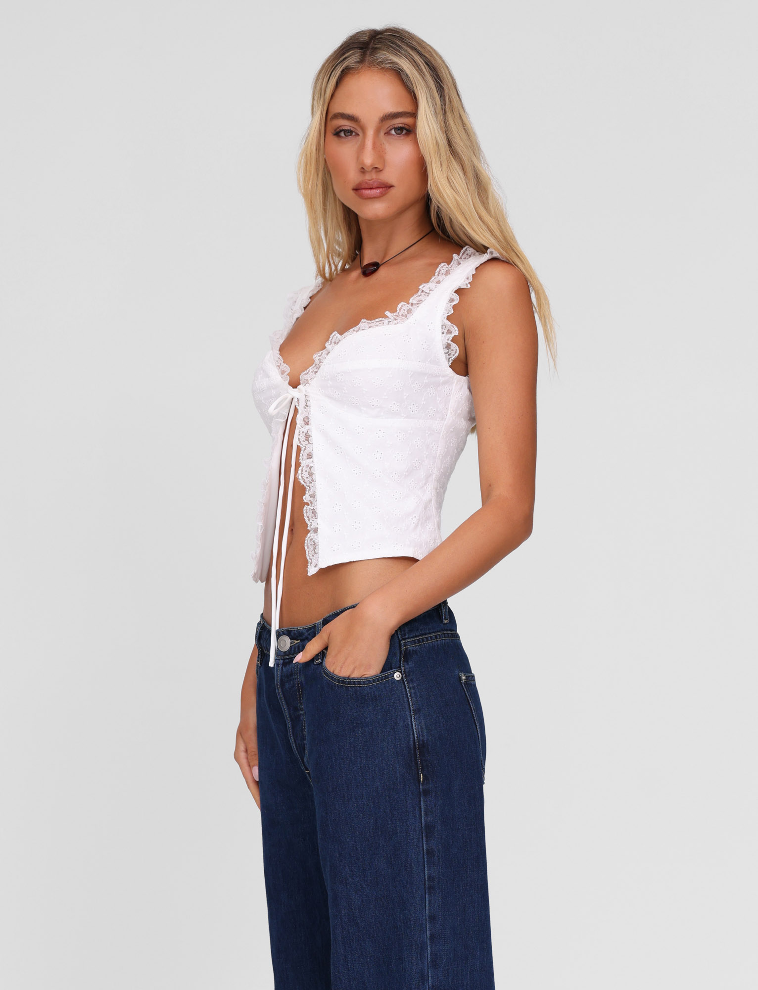A woman with long blonde hair wears the TIGERMIST KYANA TOP in white—a broderie camisole with a sweetheart bust and lace trim—paired with high-waisted blue jeans, standing against a plain light gray background.