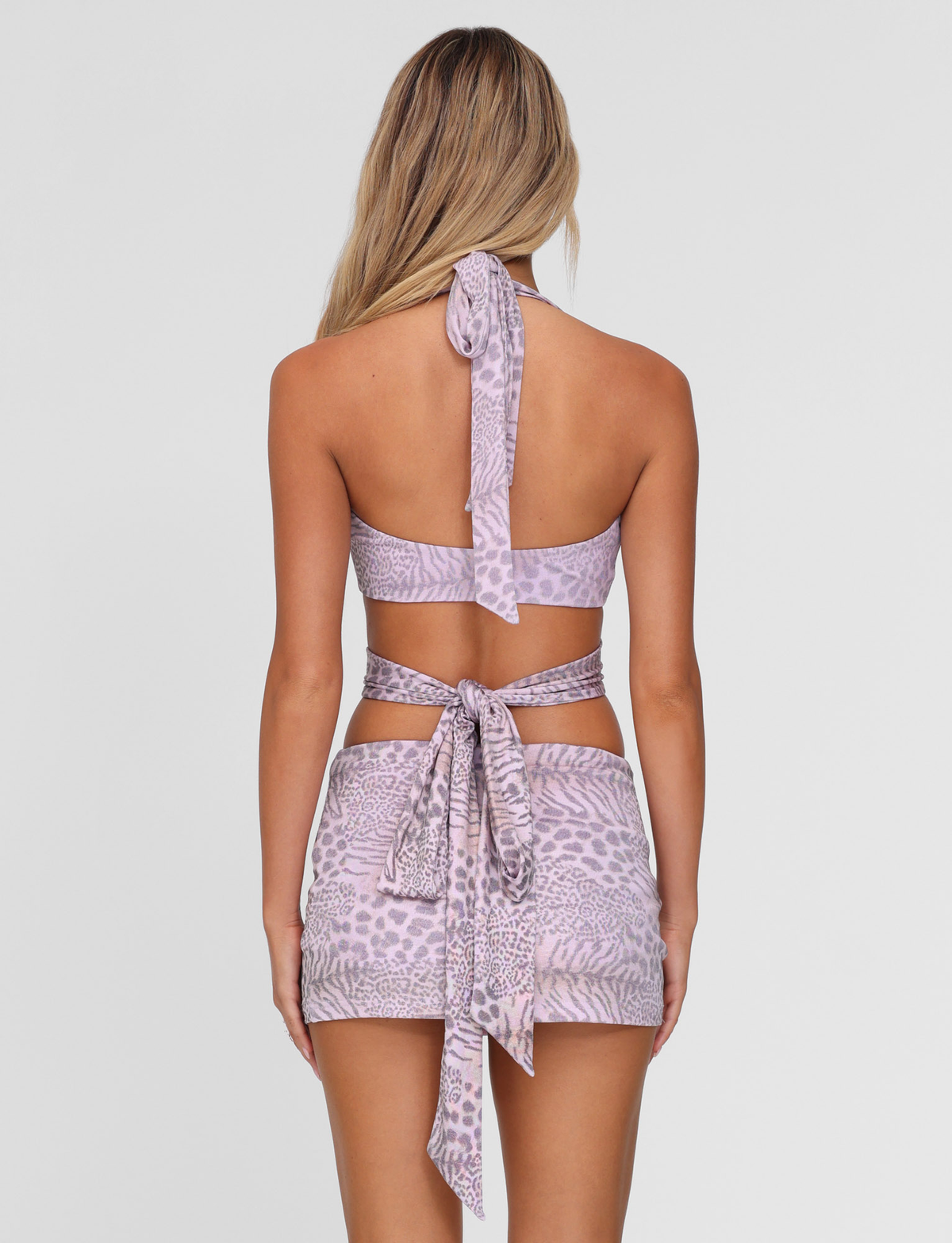 A woman with long, blonde hair is seen from the back wearing the TIGERMIST EVANGELINE SKIRT in pale pink tiger print, styled as a matching halter top and mini skirt set, featuring tie details and a bodycon fit against a white background.