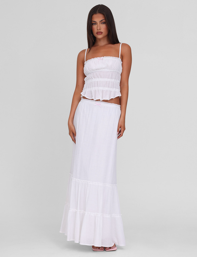 A woman with long, straight hair and a neutral expression stands against a plain background wearing the TIGERMIST ASHLYNN TOP in white. The top features adjustable straps and a tiered design, paired with a flowing white skirt. Her left hand rests on her hip.